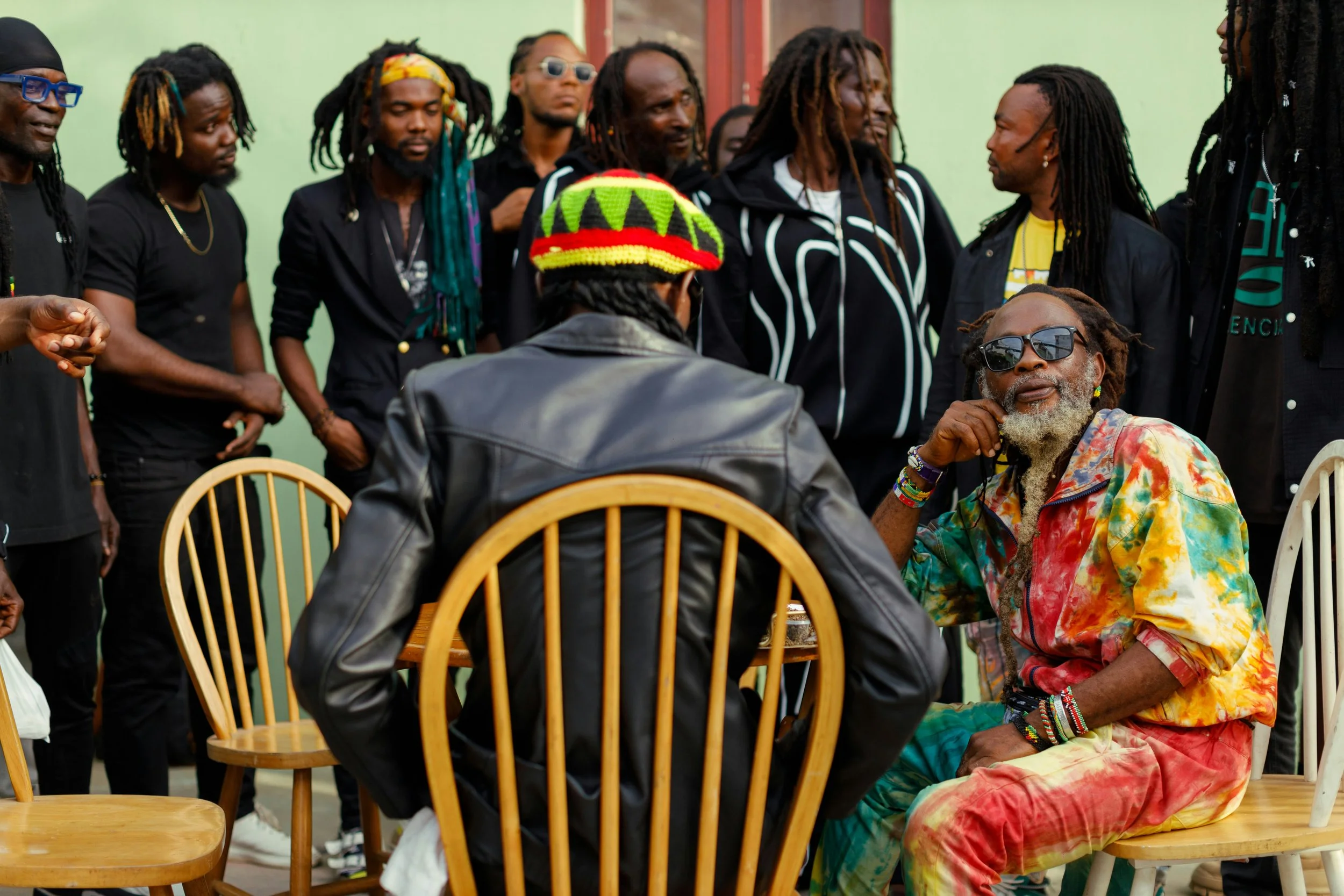Groupe de personnes avec deux hommes assis au centre, l'un portant un chapeau multicolore et un manteau en cuir, l'autre portant des vêtements colorés et des lunettes de soleil, entourés d'autres individus en arrière-plan. Le décor est un intérieur avec un mur vert pâle.