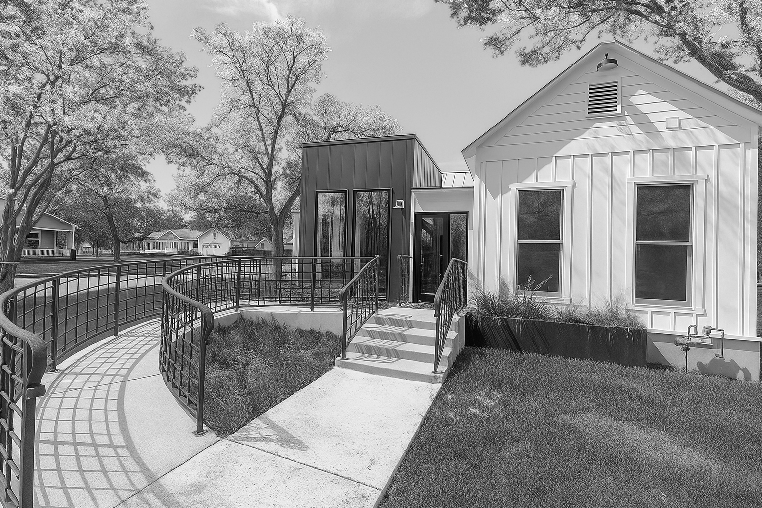 A modern house with a curved ramp and stairs leading to the entrance, featuring two large windows, a small front lawn, and a neighboring house with large trees in a suburban neighborhood.