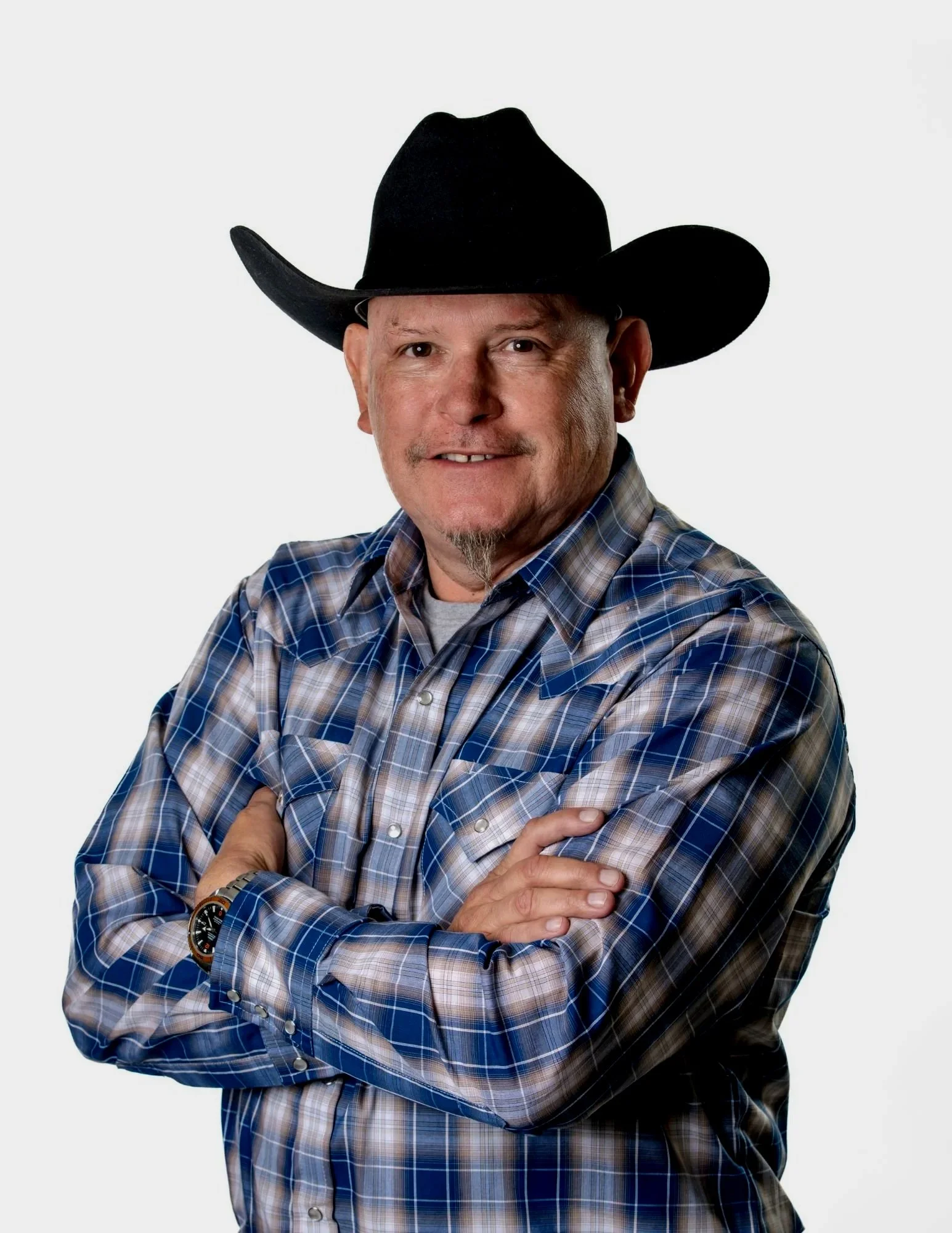 Man wearing a black cowboy hat, plaid shirt, and a watch with crossed arms, smiling against a plain white background.