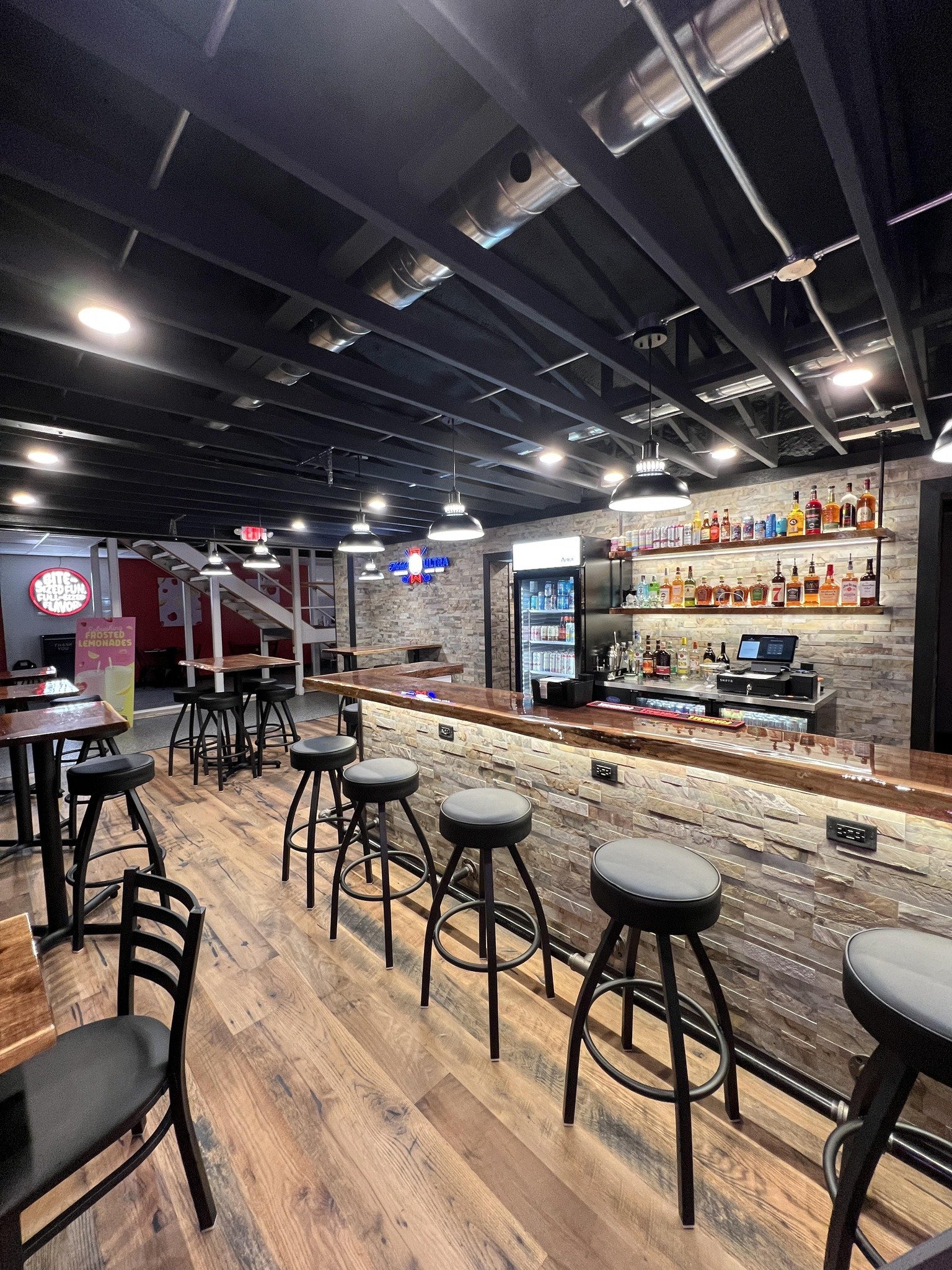 Empty bar with black stools, a wooden counter, and shelves with various liquor bottles, set in a warmly lit, modern interior.