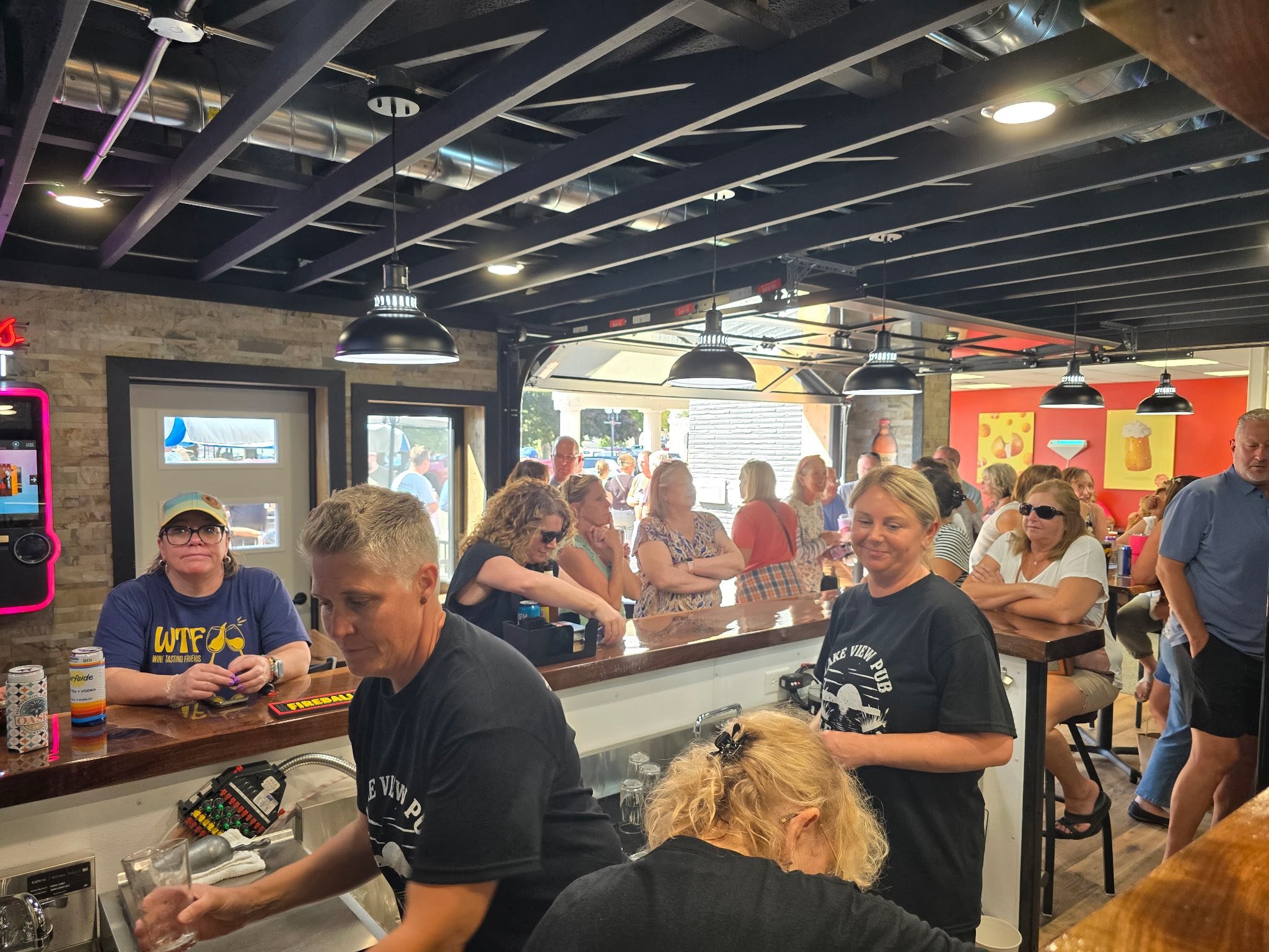 People inside a busy bar or restaurant, some standing at the counter, others sitting at tables, with bright colorful decor and hanging light fixtures.