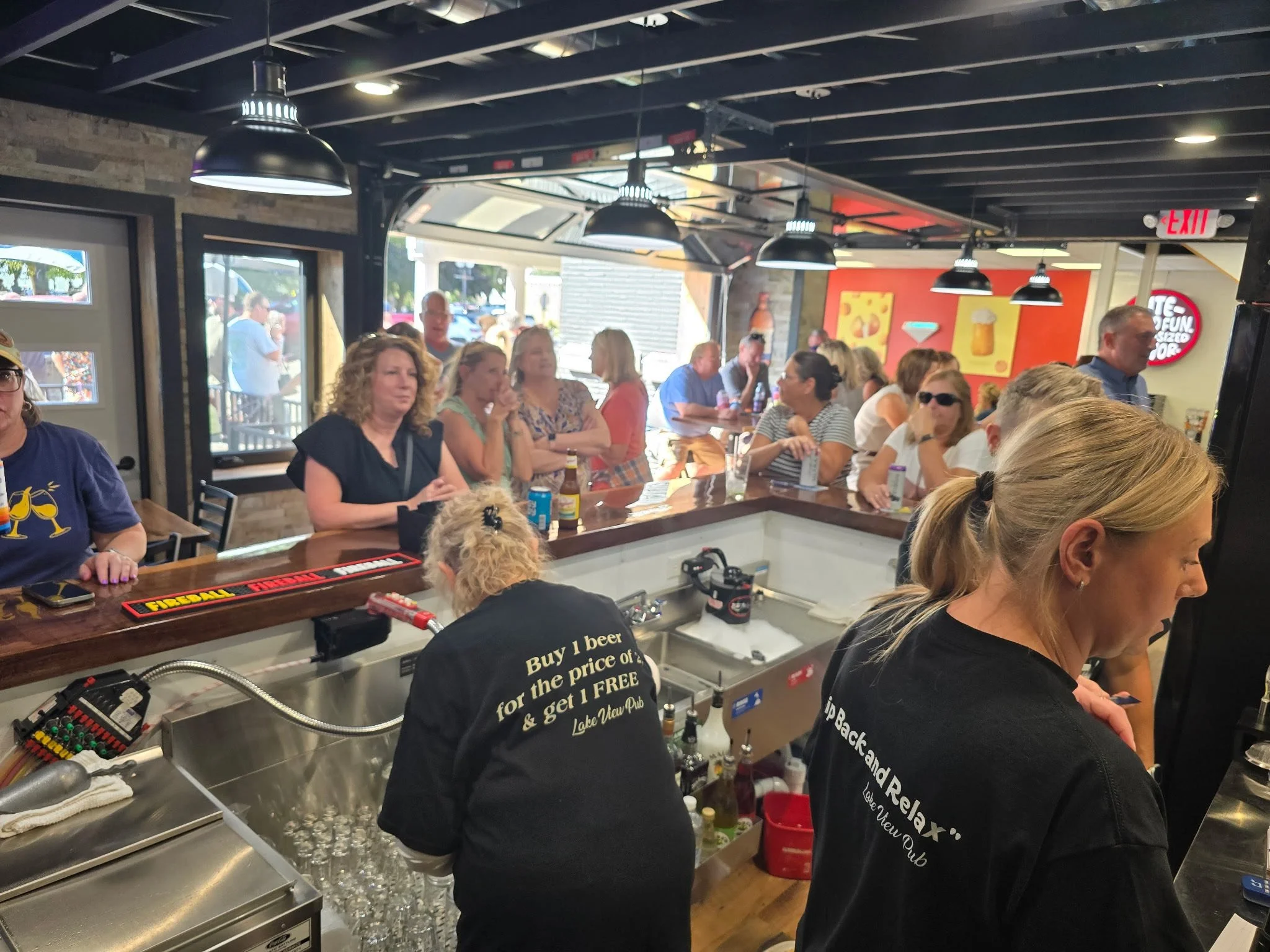 A busy bar with patrons seated and standing, bartender preparing drinks, and a window showing people outside, inside a casual restaurant or pub.