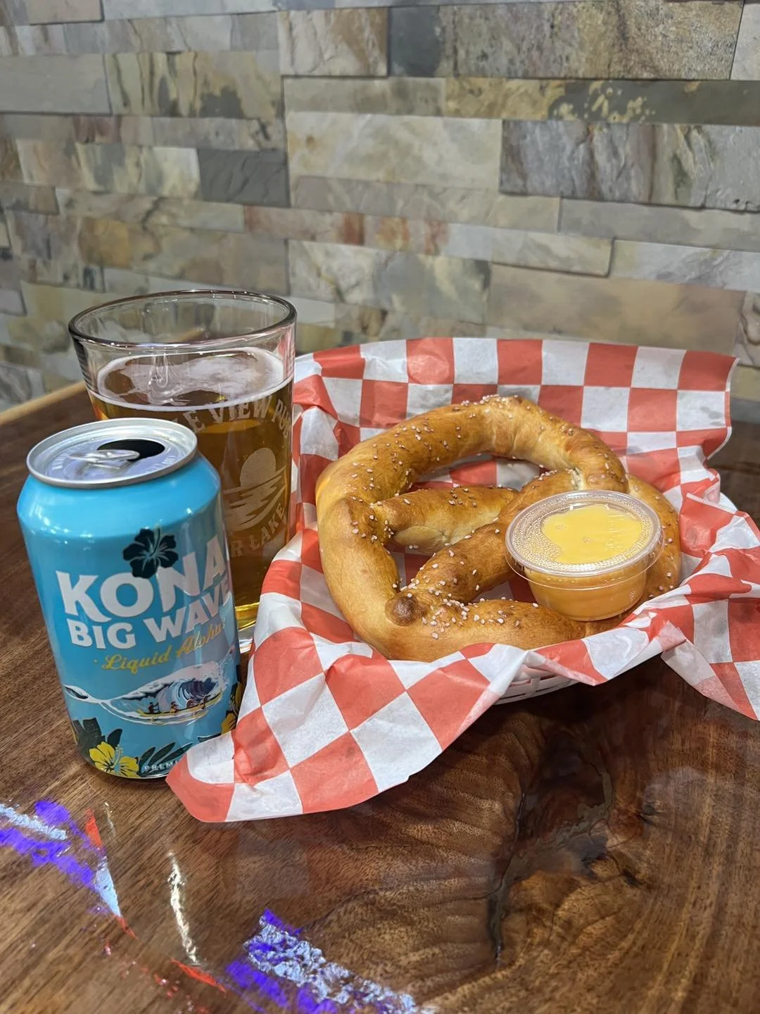 A basket with a pretzel, a small container of cheese, a glass of beer, and a can of Kona Big Wave beer on a wooden table against a stone wall background.