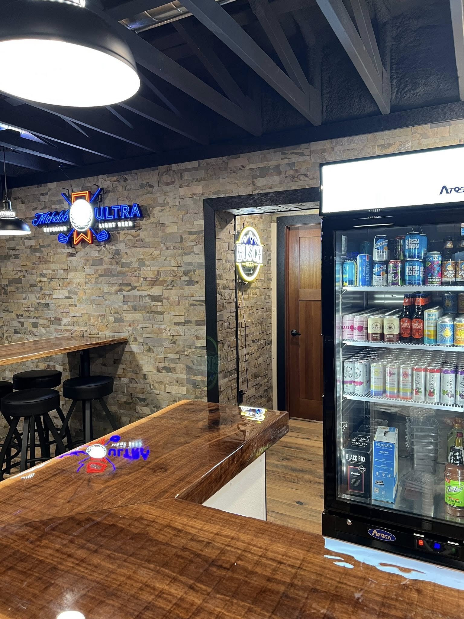 A corner of a bar or cafe with a brick wall, neon signs for Michelob Ultra and Busch beer, a refrigerated display case with drinks, a wooden counter, and black stools.