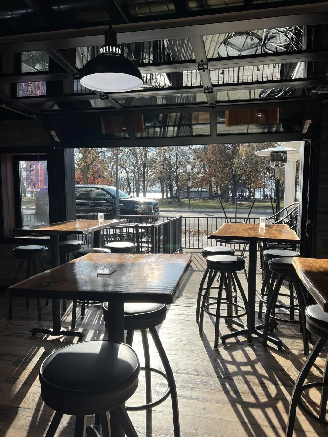 Interior of a coffee shop or restaurant with wooden tables, black bar stools, and large windows showing a view of a street with trees and parked cars outside.