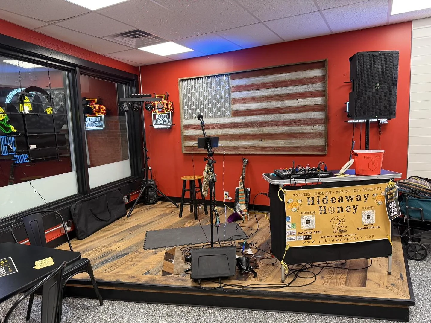 Small stage setup inside a venue with an American flag made of wood on a red wall, music equipment, and a banner reading "Hideaway Honey".