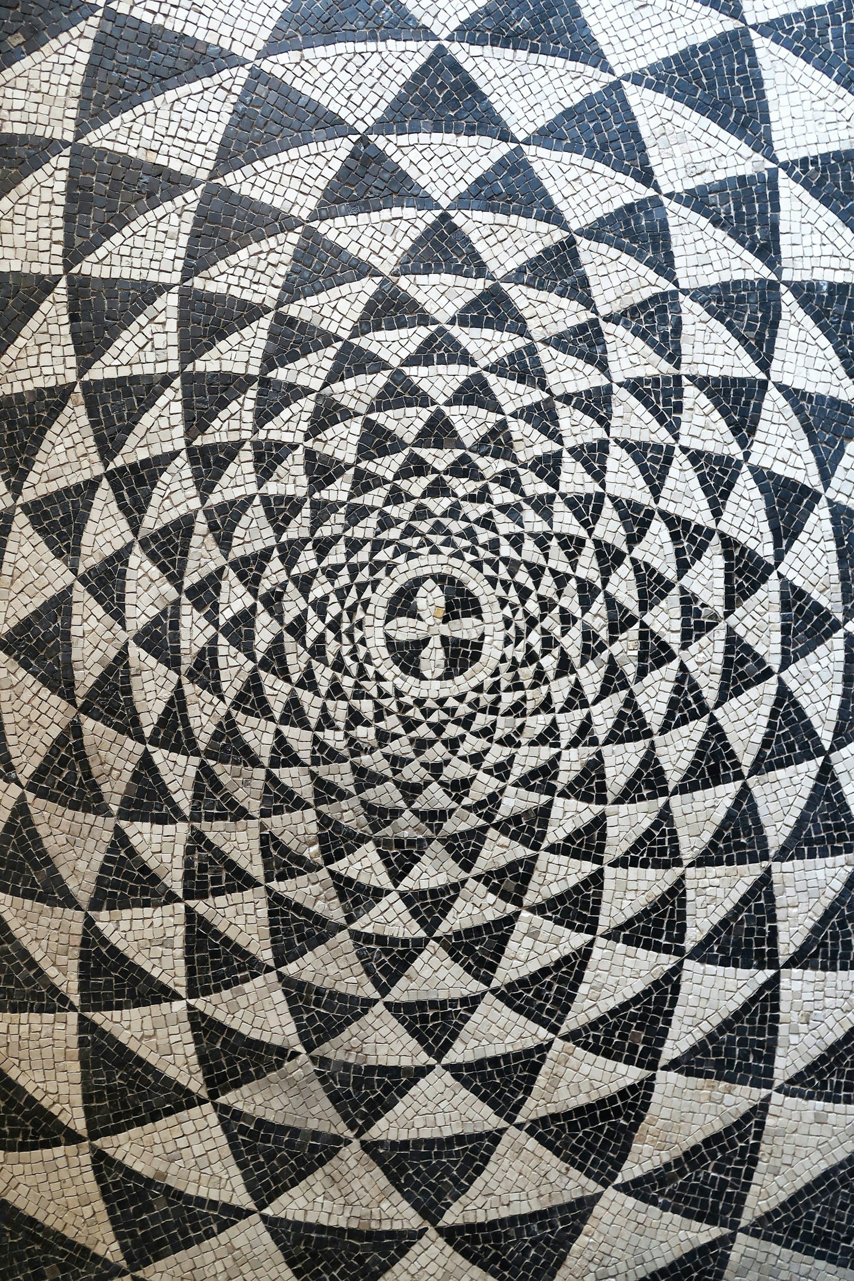 A black and white mosaic with a geometric spiral pattern, consisting of triangles, diamonds, and circles.