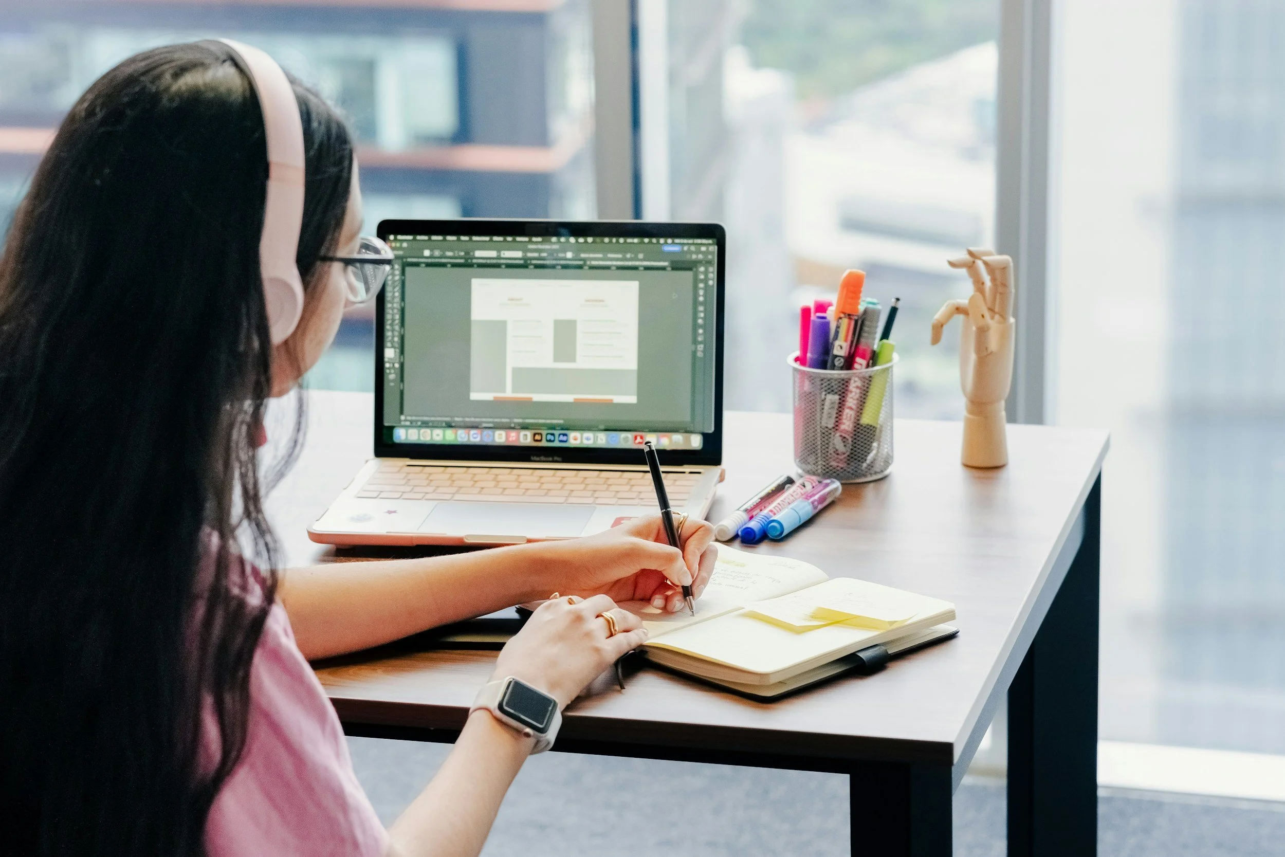 A woman with long dark hair, wearing glasses and headphones, working at a desk with a laptop, open notebook, and various pens and markers, in front of a large window.