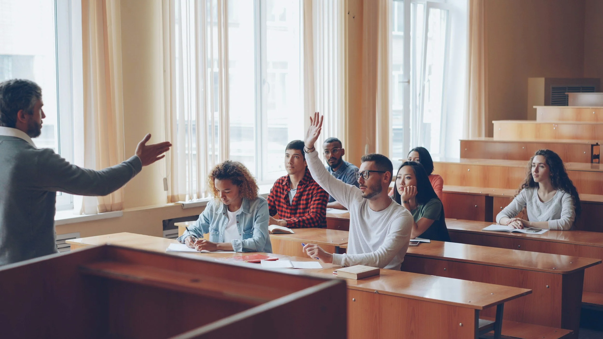 A classroom filled with students sitting at wooden desks, with one student raising their hand to ask a question or participate. The teacher, standing at the front, gestures as they speak. The classroom has large windows with curtains, allowing natural light to fill the room.