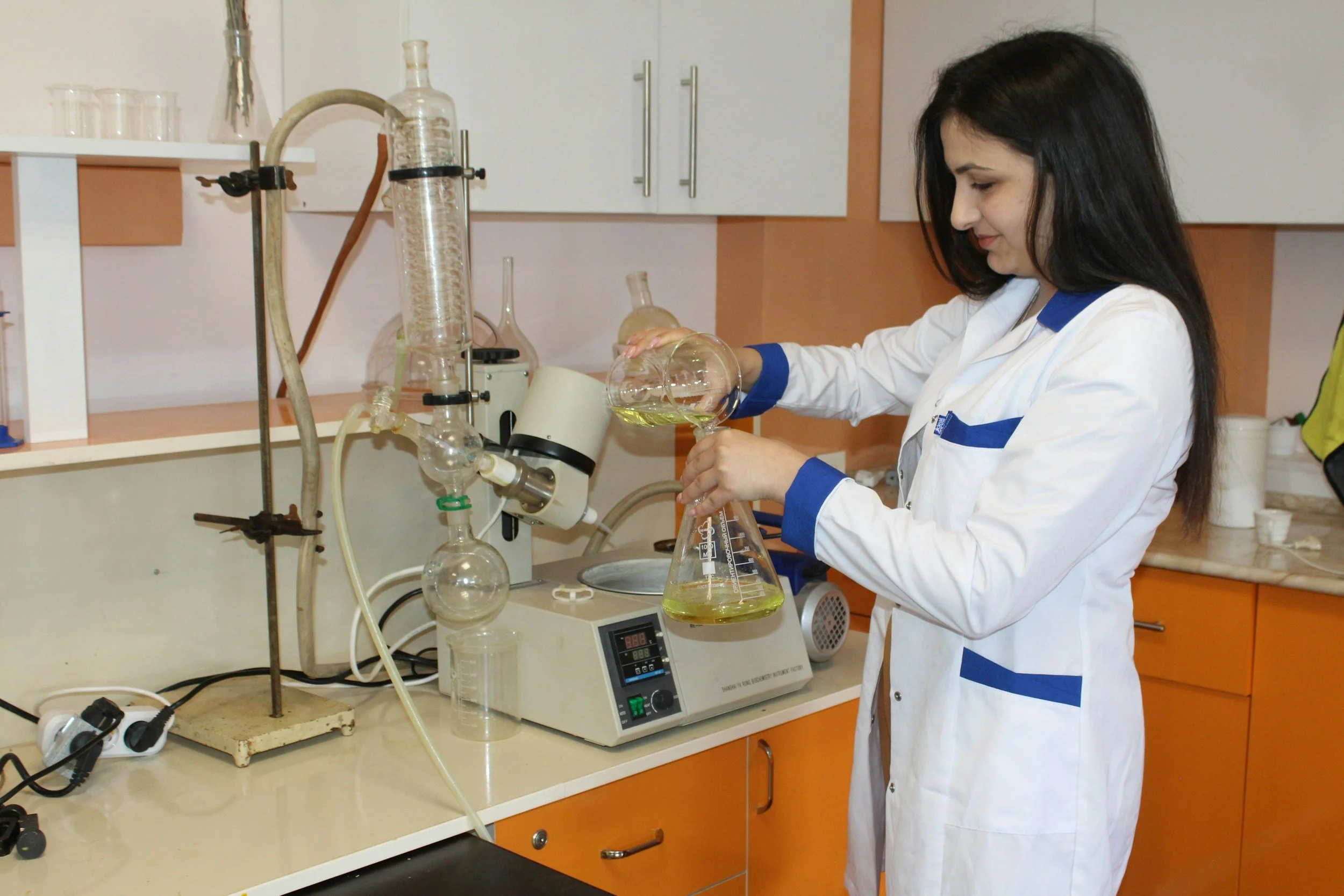 Young woman in a white lab coat pouring liquid into a beaker in a laboratory with scientific glassware and equipment on the counter.