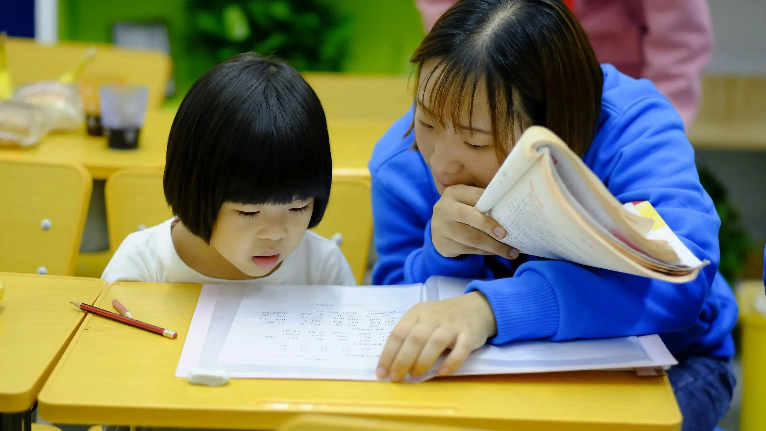 A teacher helps a young girl with her homework at a yellow desk in a classroom. The girl looks focused on her papers, while the teacher leans in closely, holding a book.