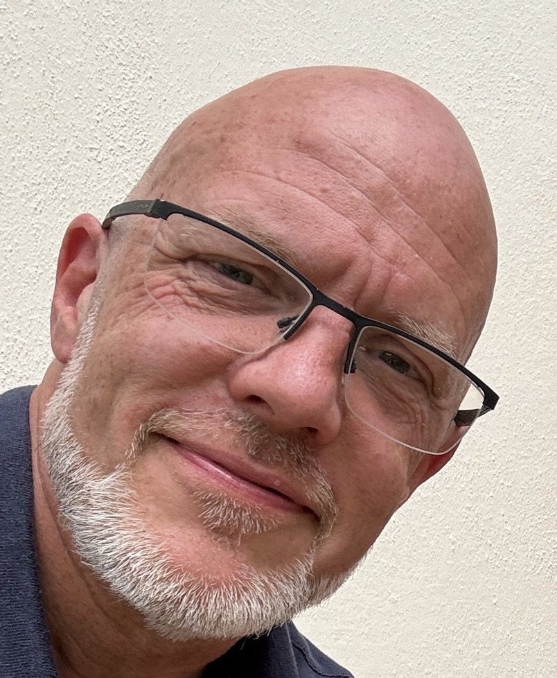 Close-up Science Tutor at home, friendly and professional smiling bald man with glasses and a white beard, wearing a dark shirt, against a textured off-white wall.