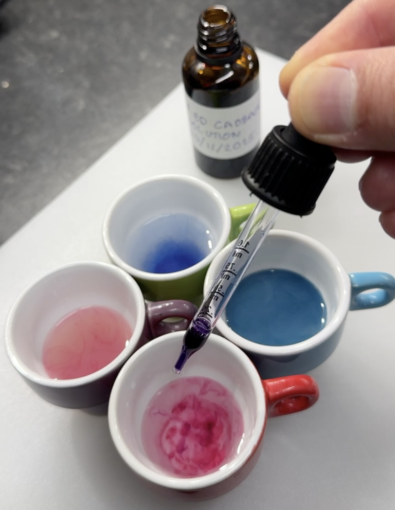 A hand holding a dropper with purple liquid, about to drop it into a small pink and white marble-effect cup. Four small cups filled with different colored liquids are on a white surface, four mugs in various colors surrounding them. A small brown bottle with a white label is in the background.