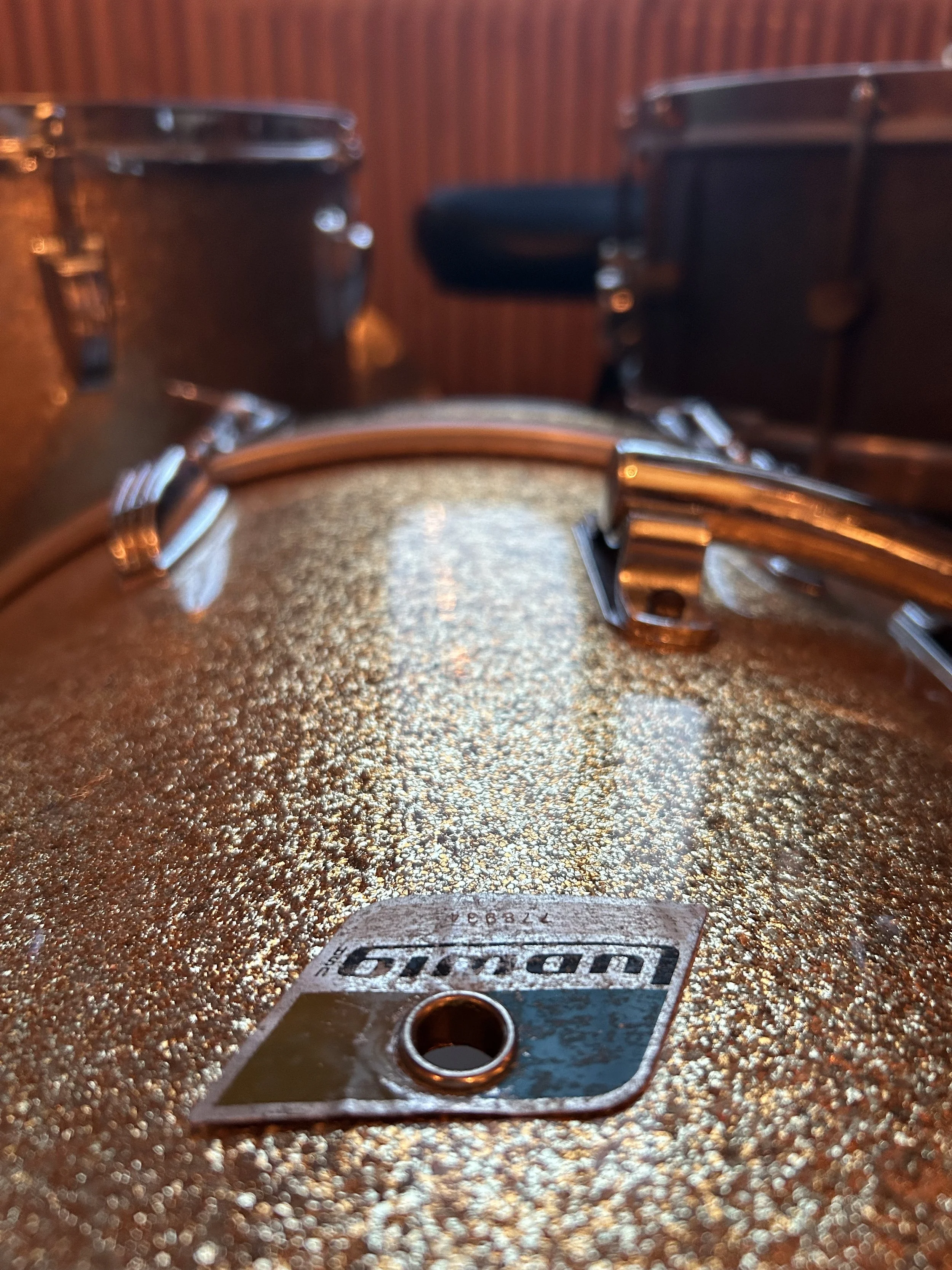 Close up of a 1970's Ludwig bass drum in champagne sparkle with blue and olive badge. In the back: Floor tom and snare drum in front of wooden panels. Golden hour, warm colors.