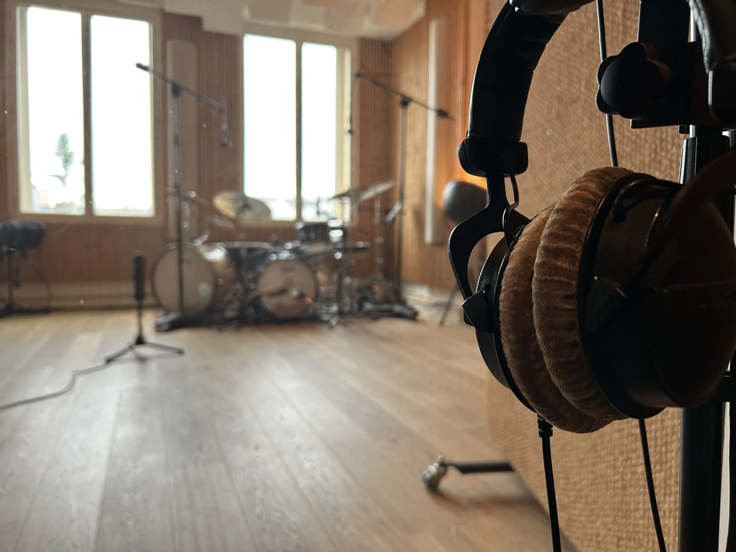 Beyerdynamic headphones hanging on a mic stand in a recording studio. In the back a fully mic'ed drum kit with two bass drums. The studio has wooden panels and big windows.