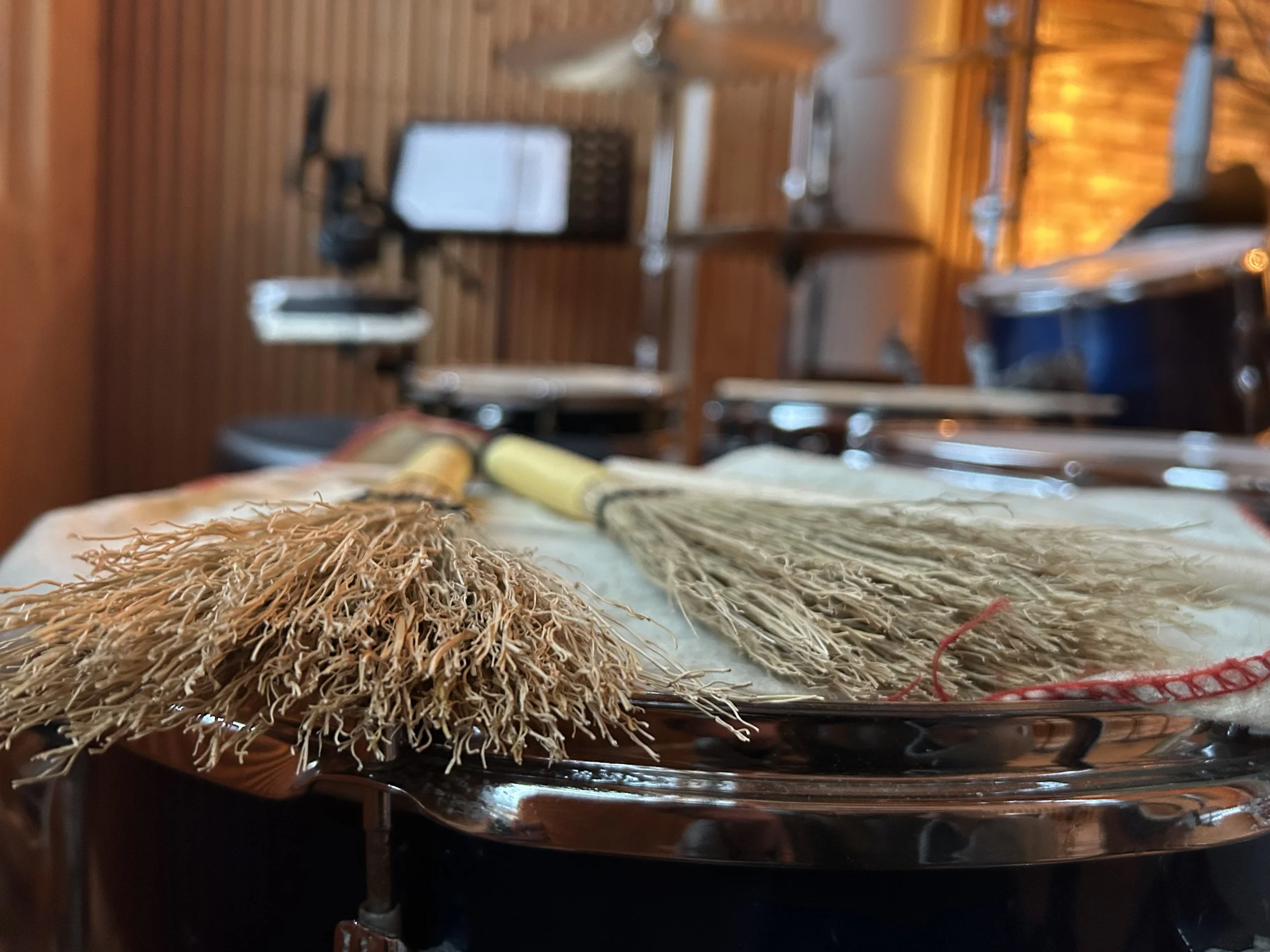A pair of corn, straw drum brushes laying on a floor tom. In the background the rest of the blue shimmer Premier drum kit with several microphones in a recording studio with wooden panels and a wall made out of red bricks.