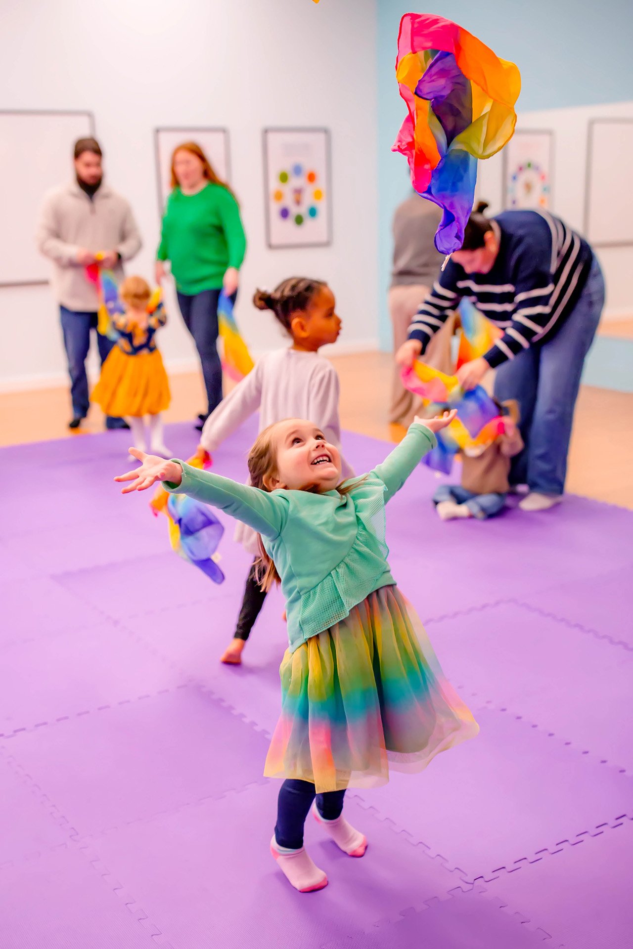 Children playing with rainbow-colored fabric streamers in a room with purple mats and framed artwork on the walls.