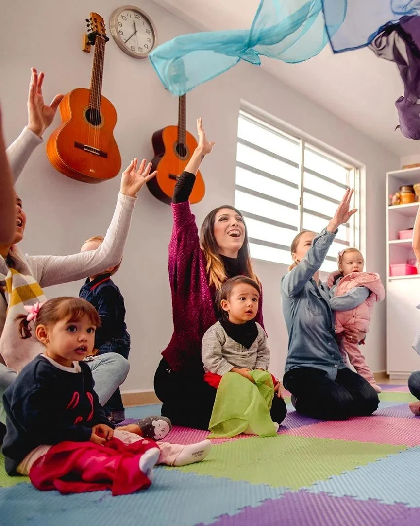 Children and a woman in a music class sitting on colorful mats, reaching towards colorful scarves in the air. Guitars hanging on the wall and a clock are visible in the background.
