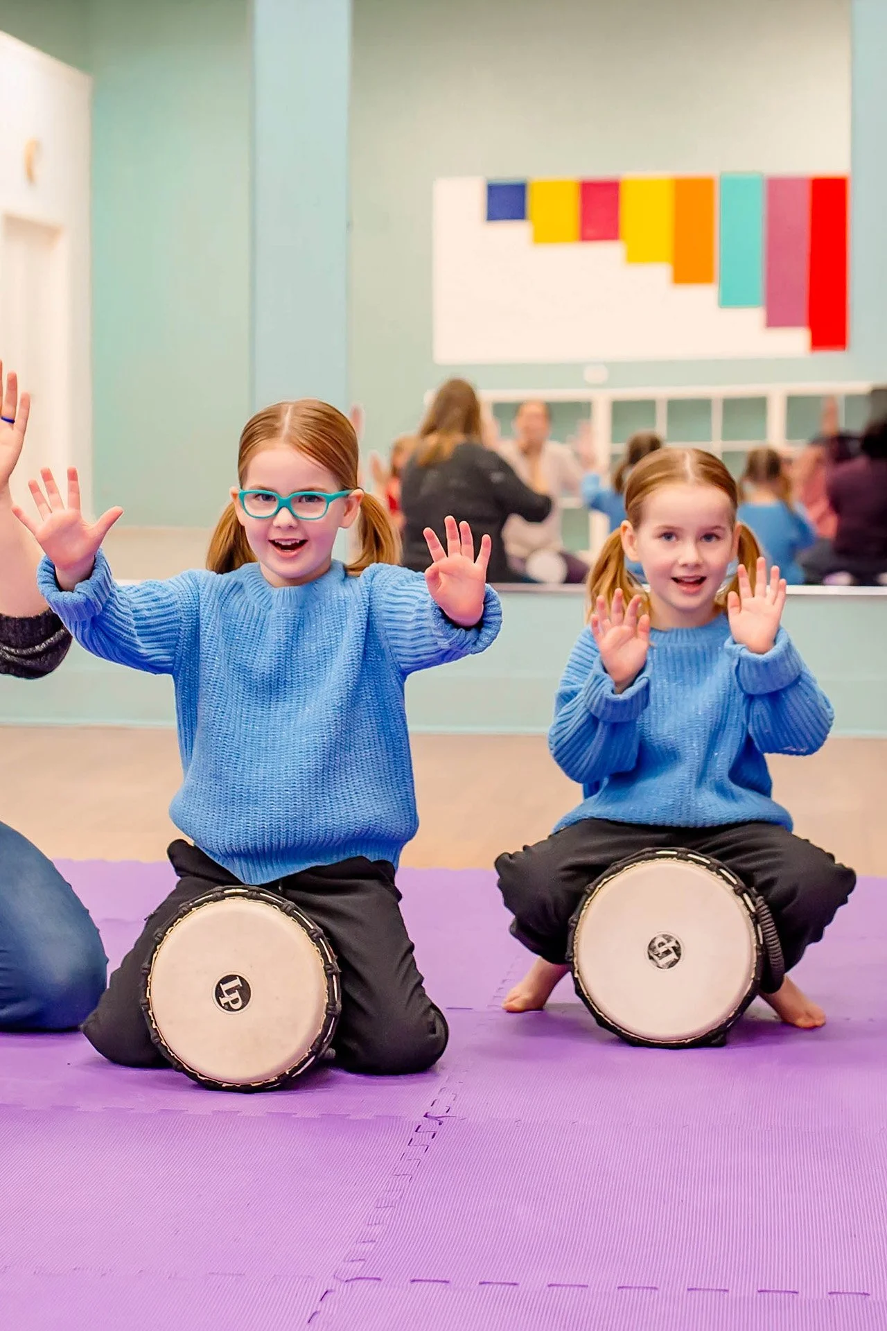 Two young girls with pigtails and glasses, wearing blue sweaters, sitting on purple mats, playing drums and raising their hands, in a colorful indoor setting with an audience in the background.