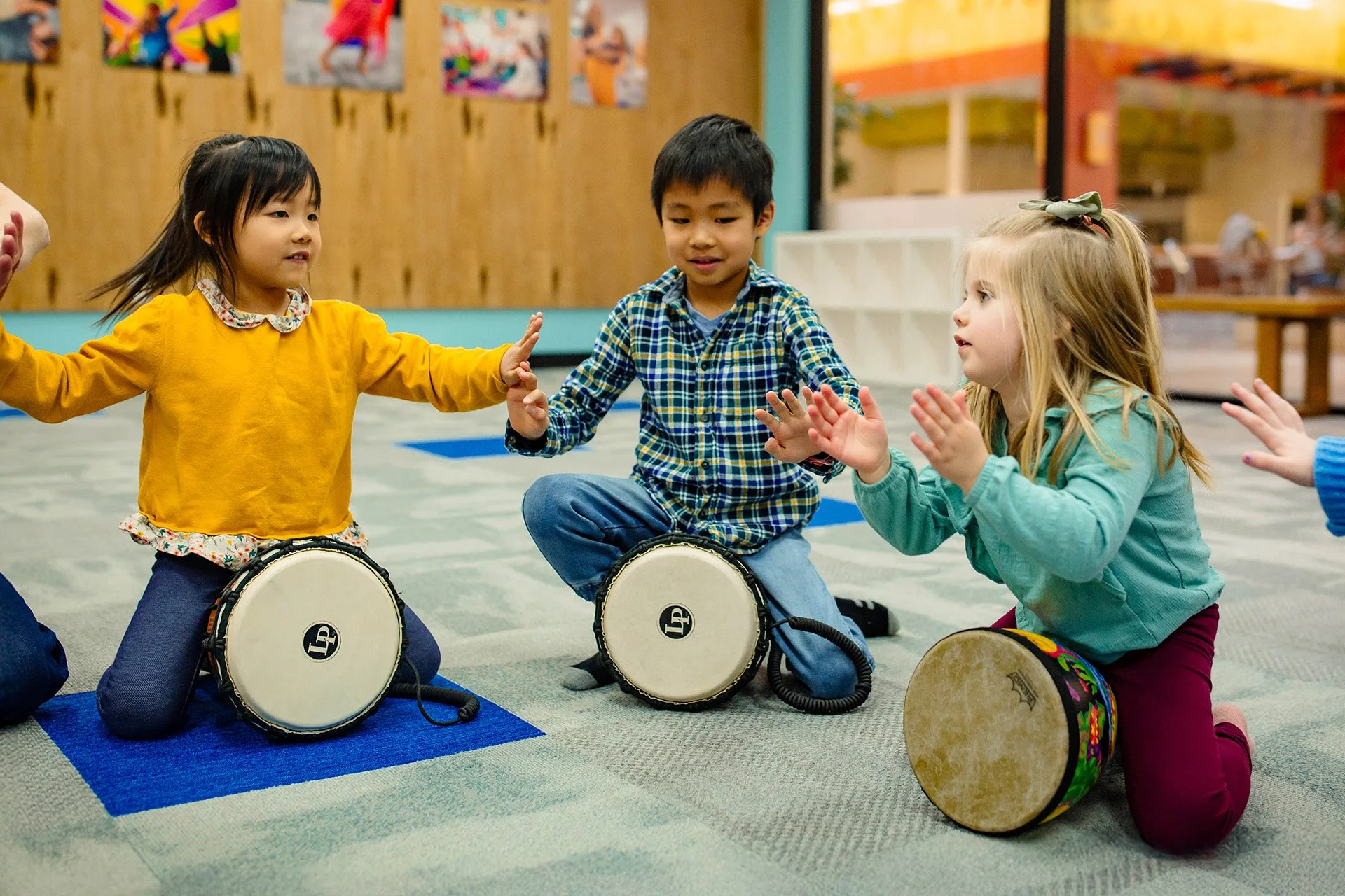 Three children sitting in a circle playing small drums in a classroom.