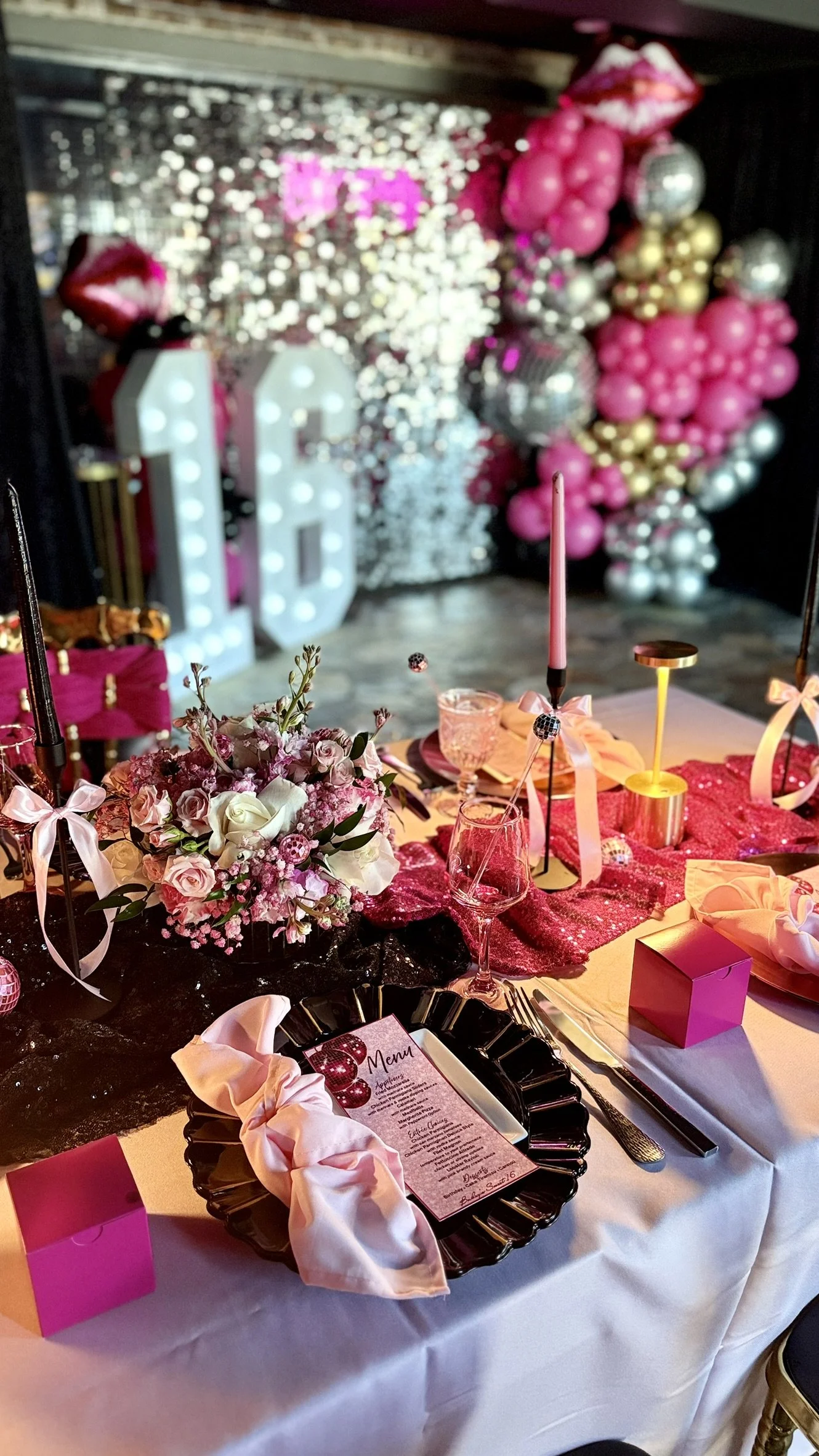 Decorated party table with pink, white, and gold accents, floral centerpiece, pink candles, and pink boxes with balloons in the background. Shimmer wall, Jumbo LED Numbers, balloon garland, LED sign
