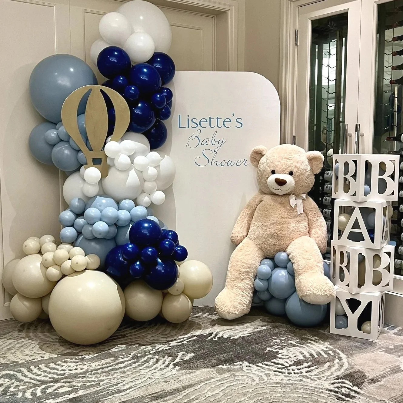 Balloon arrangement in blue, white, and cream hues with a hot air balloon cutout, a white sign reading "Lisette's Baby Shower," a large teddy bear, and white blocks spelling "BABY" next to a wine rack.