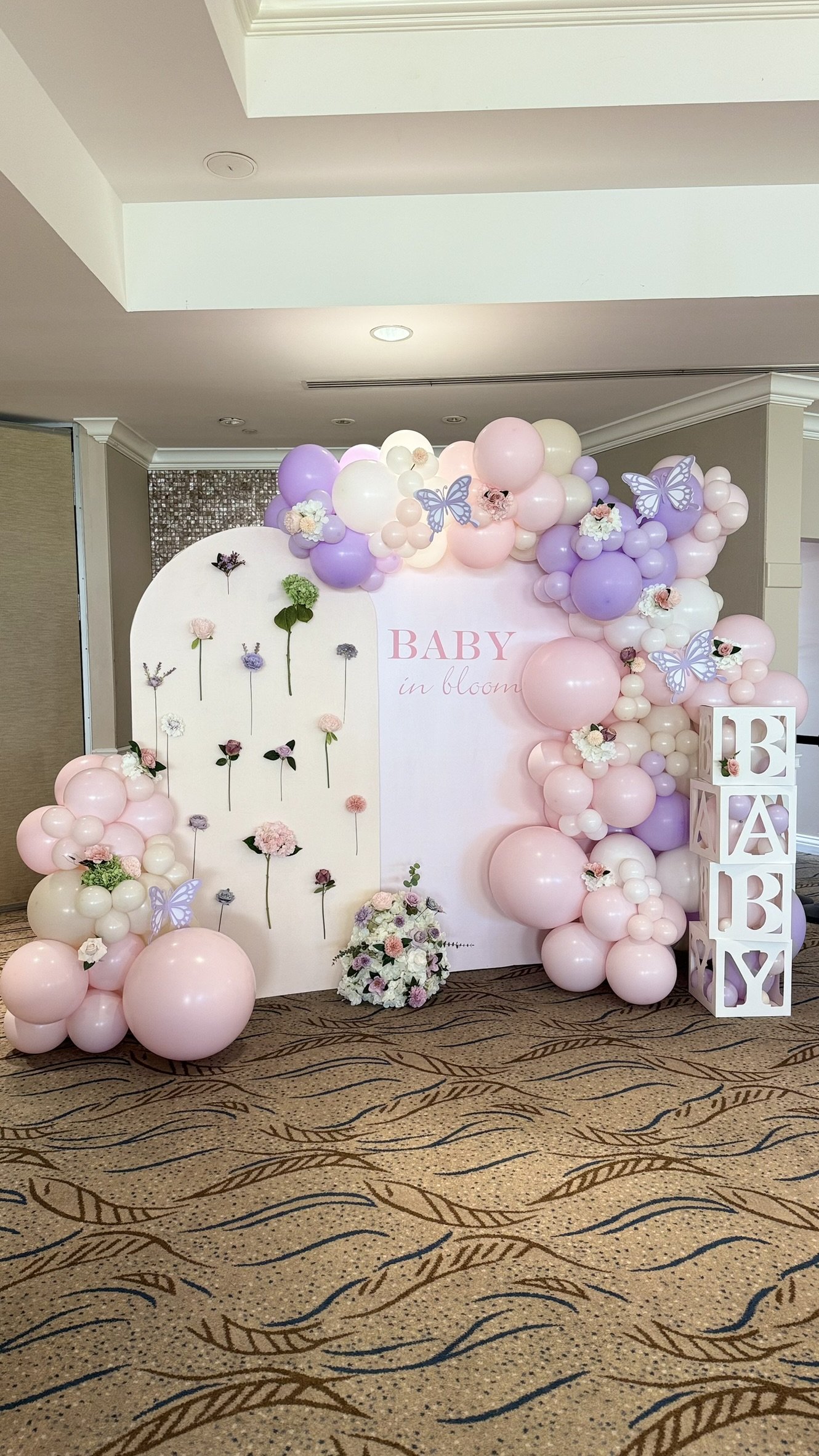 Baby shower backdrop decorated with pink, purple, and white balloons, butterflies, flowers, and a white cube display spelling 'BABY'. A white board with 'BABY in bloom' written on it, and a flower arrangement at the bottom.