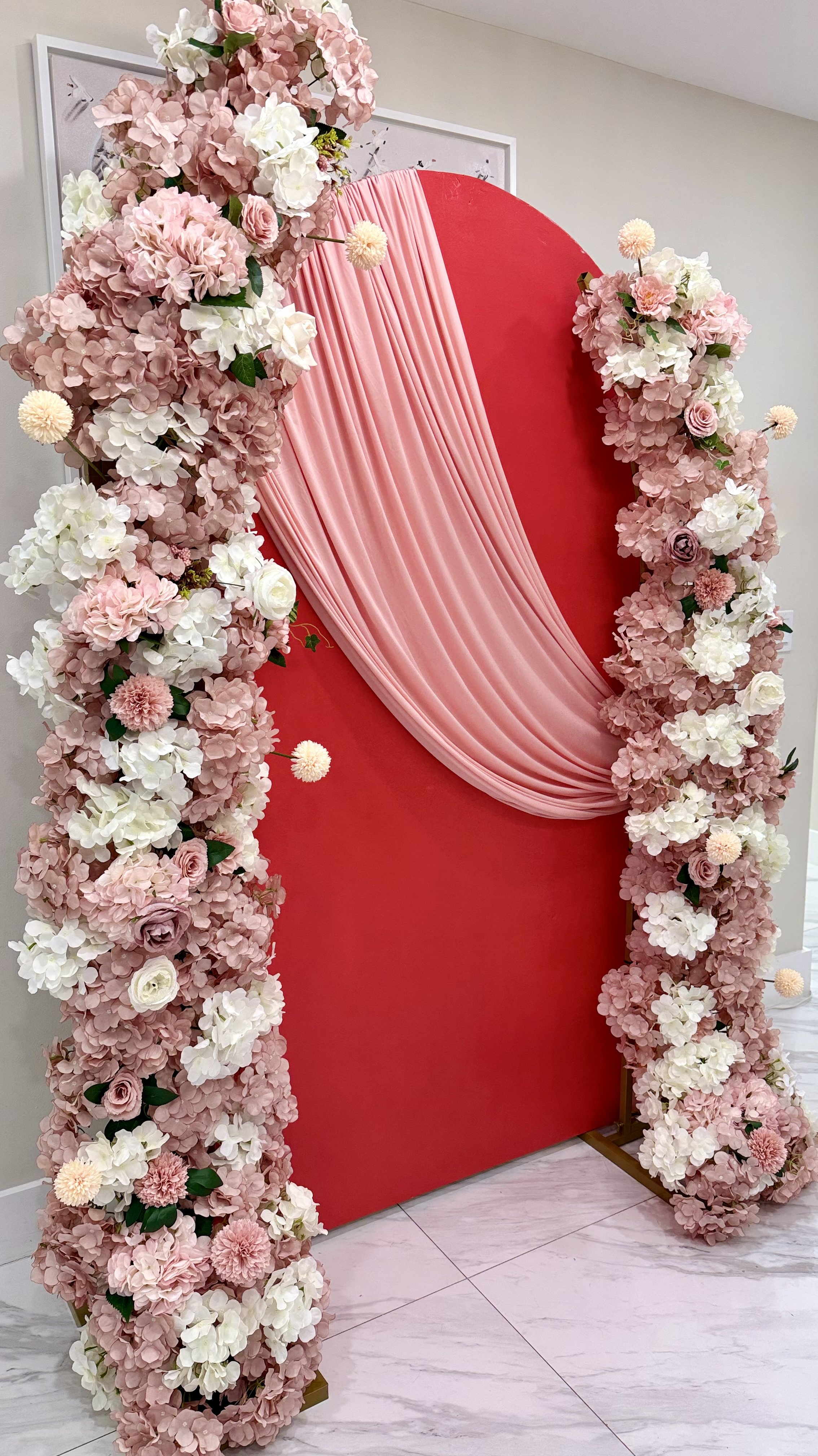A decorative backdrop with a red panel draped with pink fabric and surrounded by pink and white flowers for a special event.