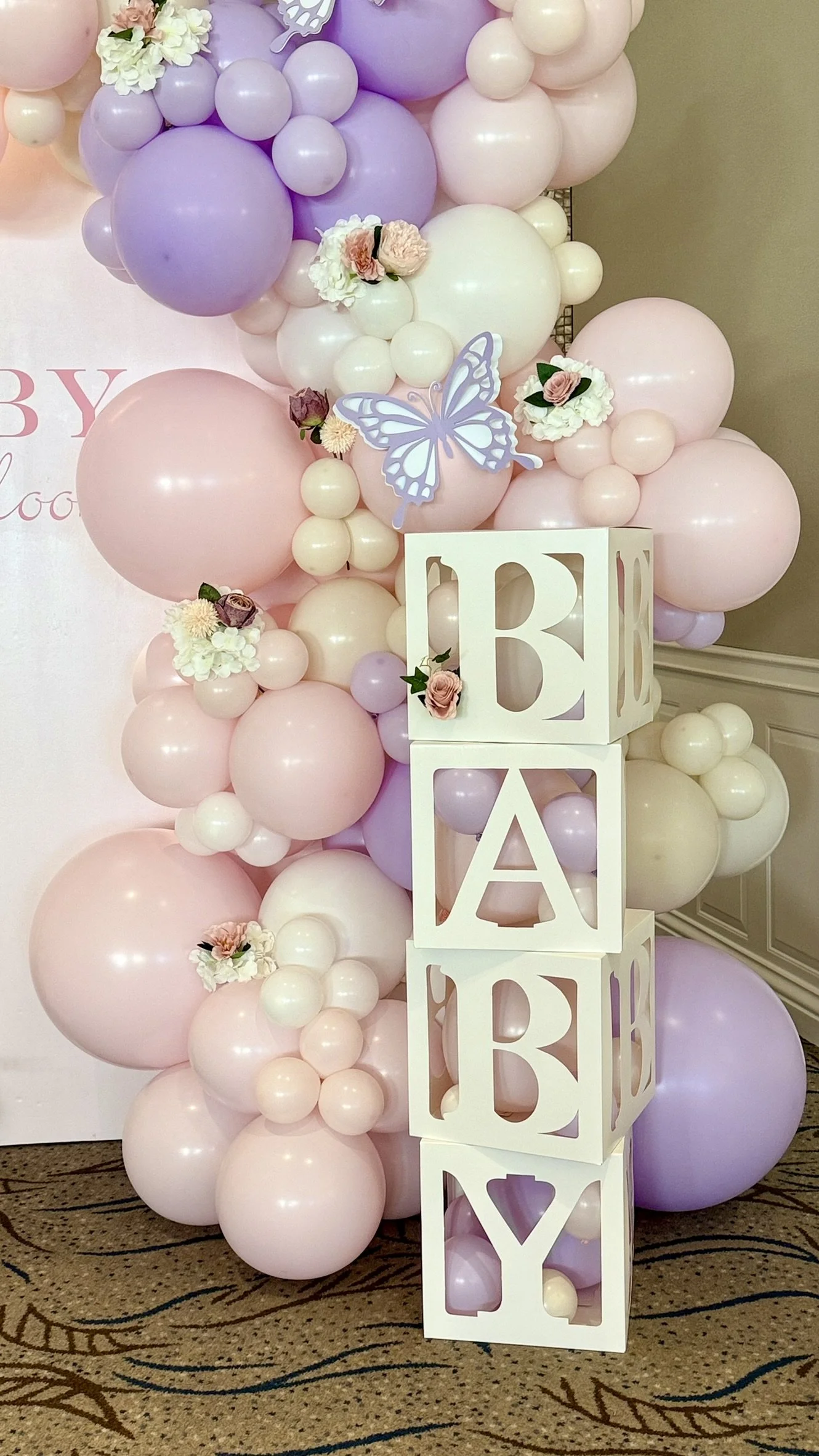 Decorative balloon arrangement in shades of pink, purple, and white, with flowers, butterfly cutouts, and white block letters spelling 'BABY'