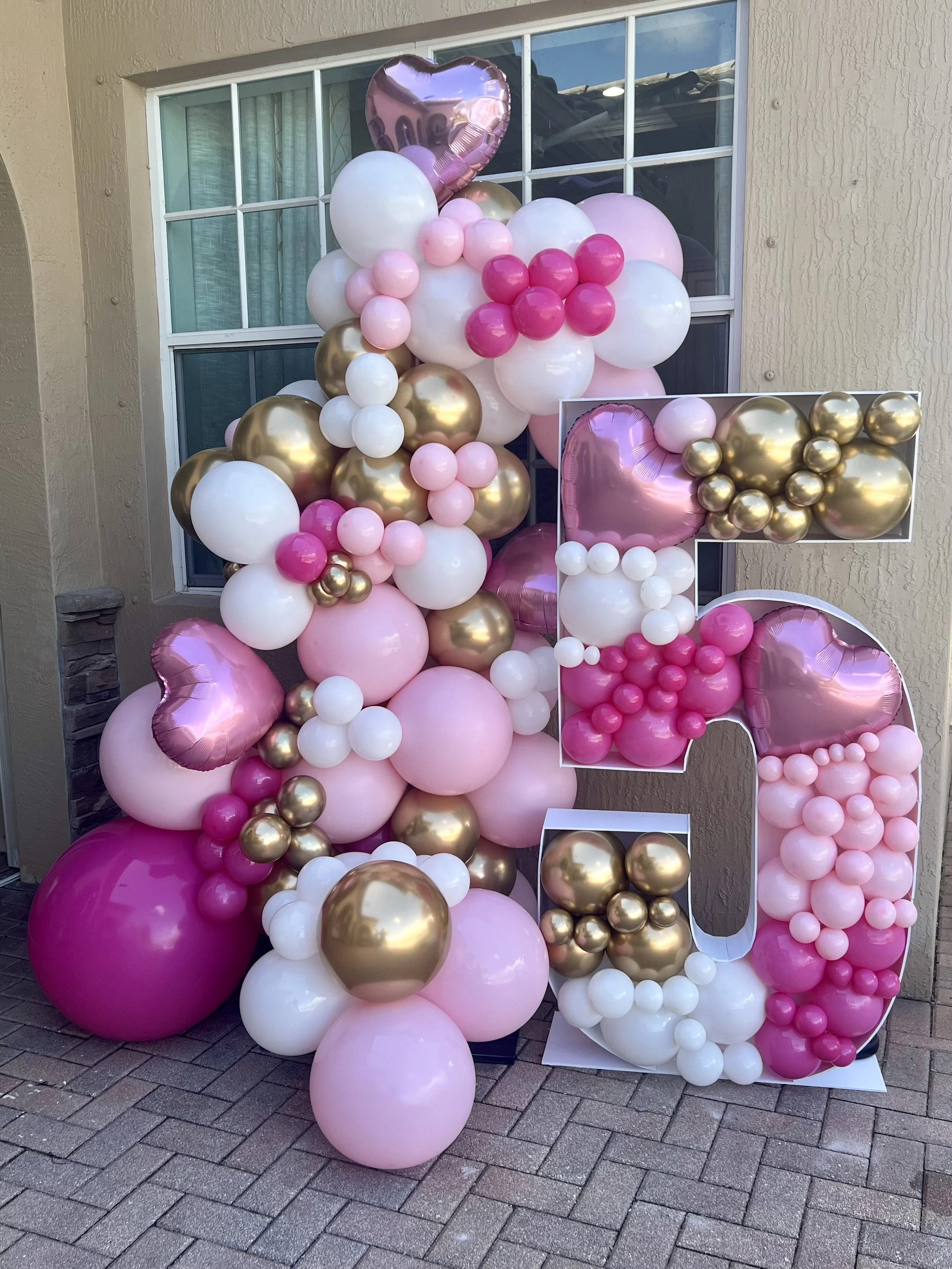 A decorative balloon display with pink, white, and gold balloons, including a large letter 'G' and heart-shaped balloons, set outside a house near a window.