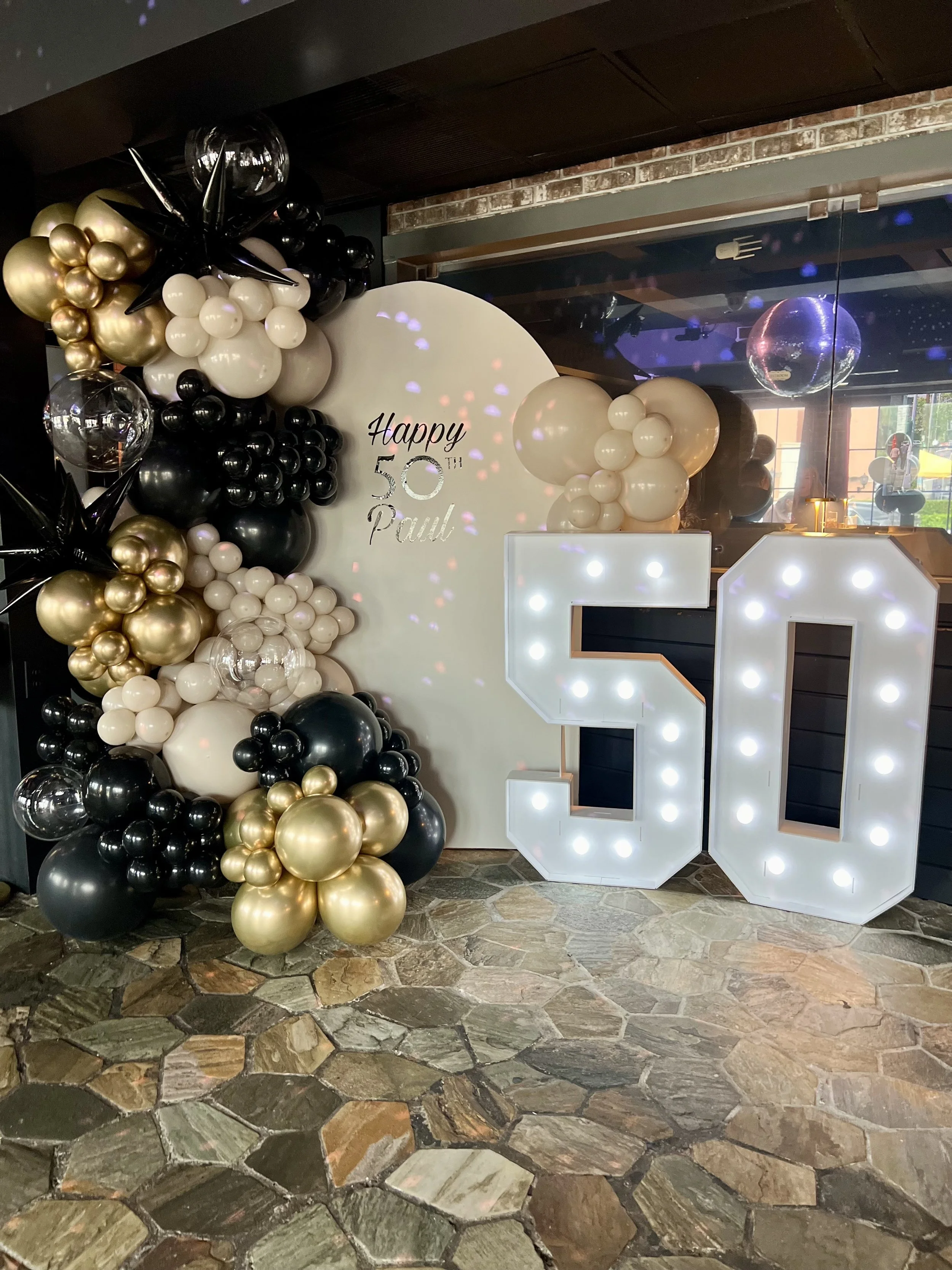 Decorative celebration display for a 50th birthday with large illuminated number 50, balloons in black, white, gold, and clear colors, a white circular backdrop with 'Happy 50th Paul' written on it, and a disco ball reflecting light, all set on a stone tile floor.
