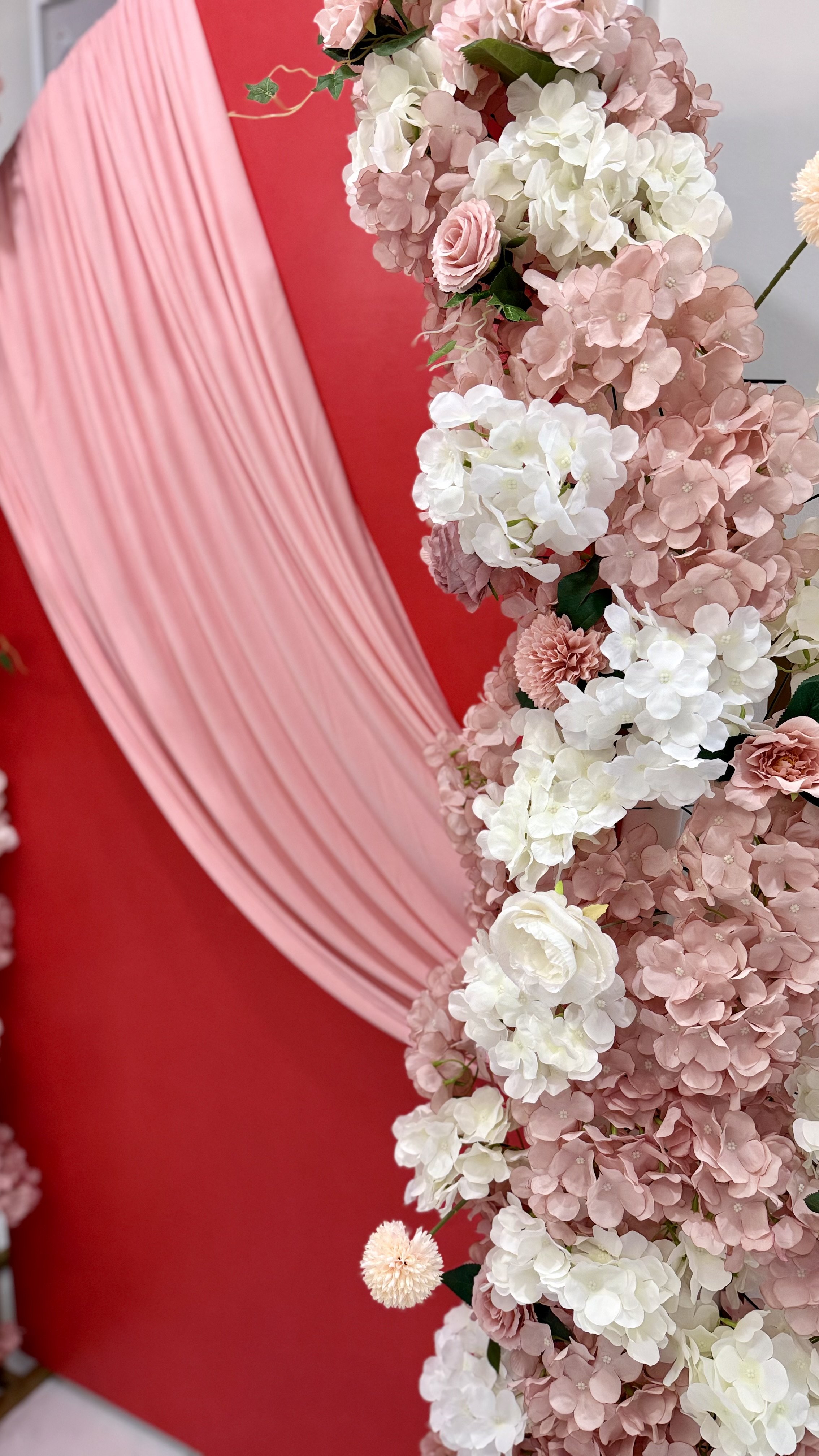 A floral arrangement of pink and white hydrangeas, roses, and carnations with a pink fabric drape against a red backdrop.