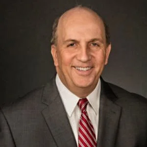 A middle-aged man in a gray suit and red striped tie, smiling, against a dark background.
