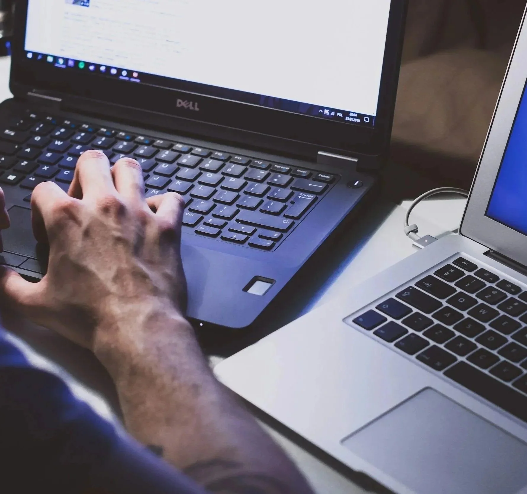 Person typing on a black Dell laptop with a silver MacBook nearby on a white table.