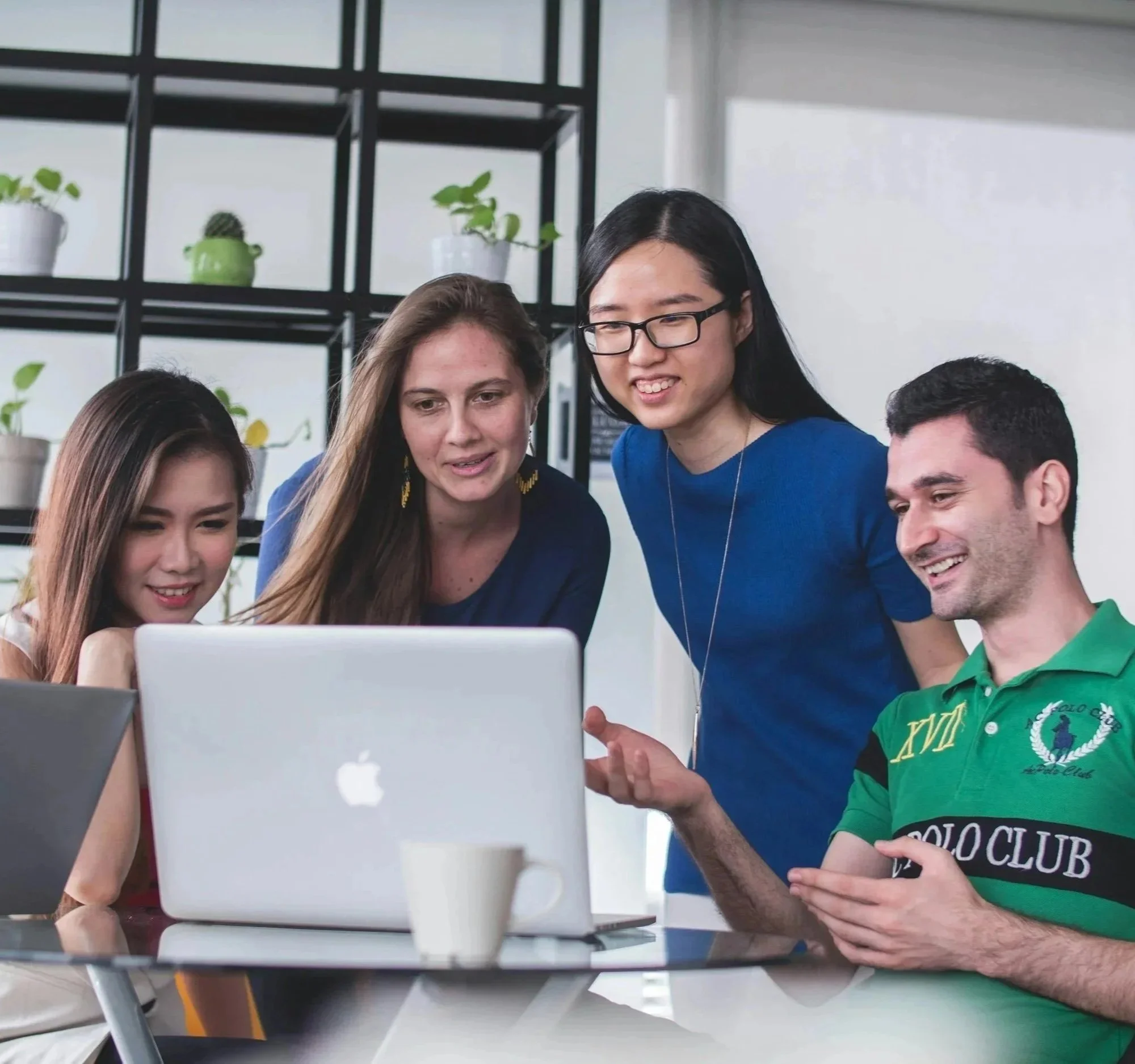 Four young adults gathered around a laptop, looking at the screen and smiling in a brightly lit room with shelves and potted plants in the background.