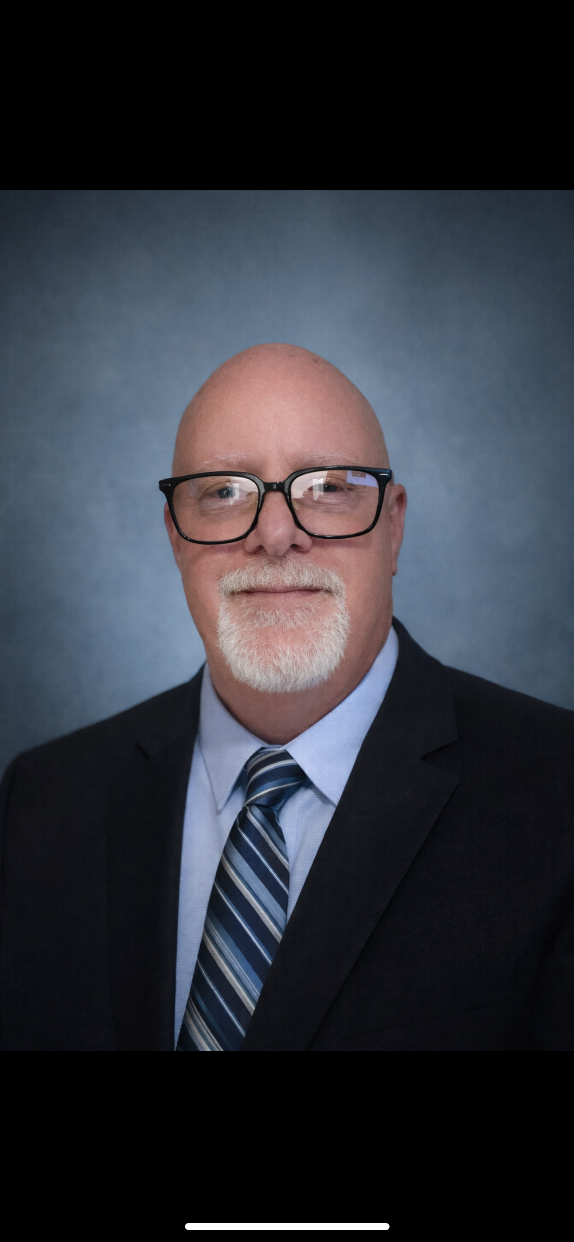 Professional headshot of a bald man with glasses, a white beard, wearing a dark suit, light blue shirt, and striped tie against a gray background.