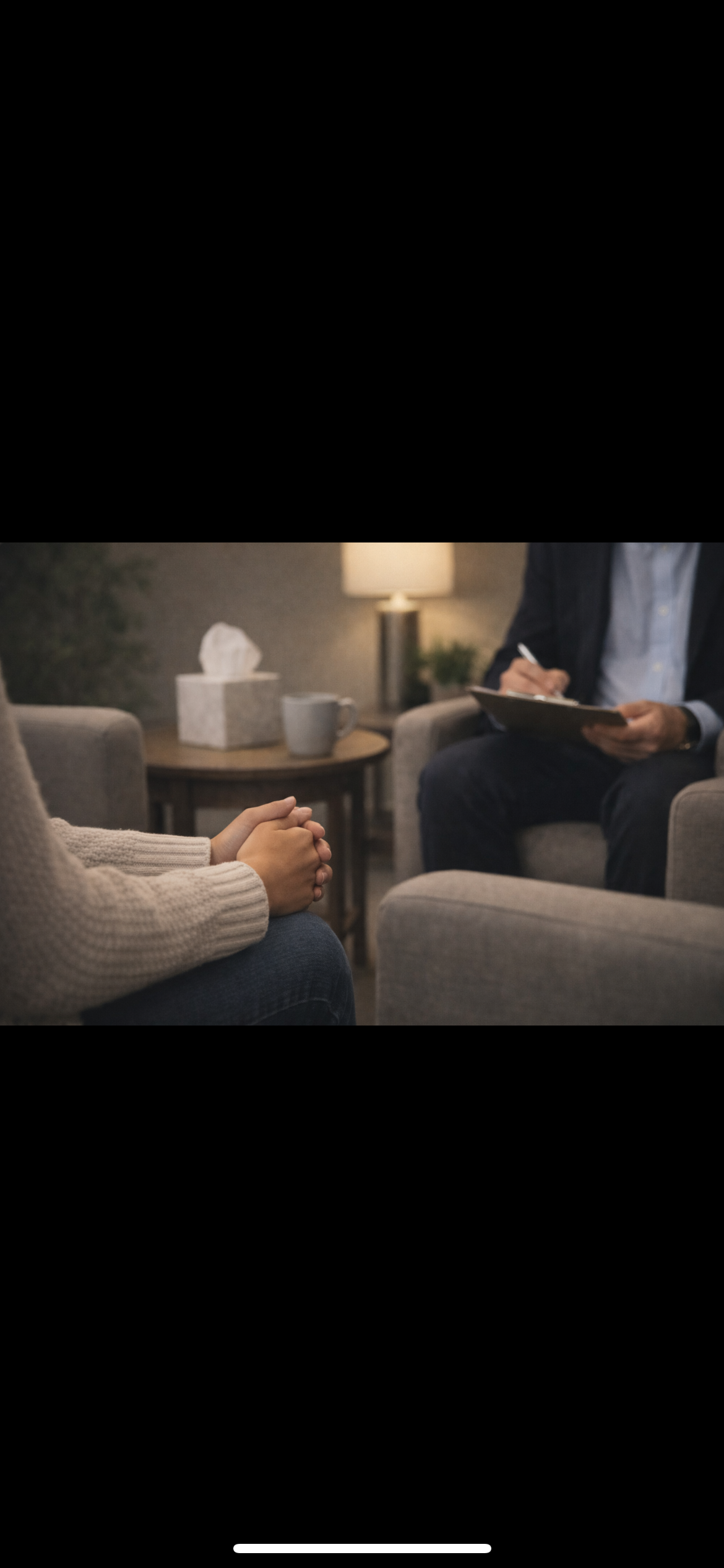 Person in a therapy session with a therapist holding a notepad, sitting in a cozy office with a lamp, tissue box, mug, and plant in the background.