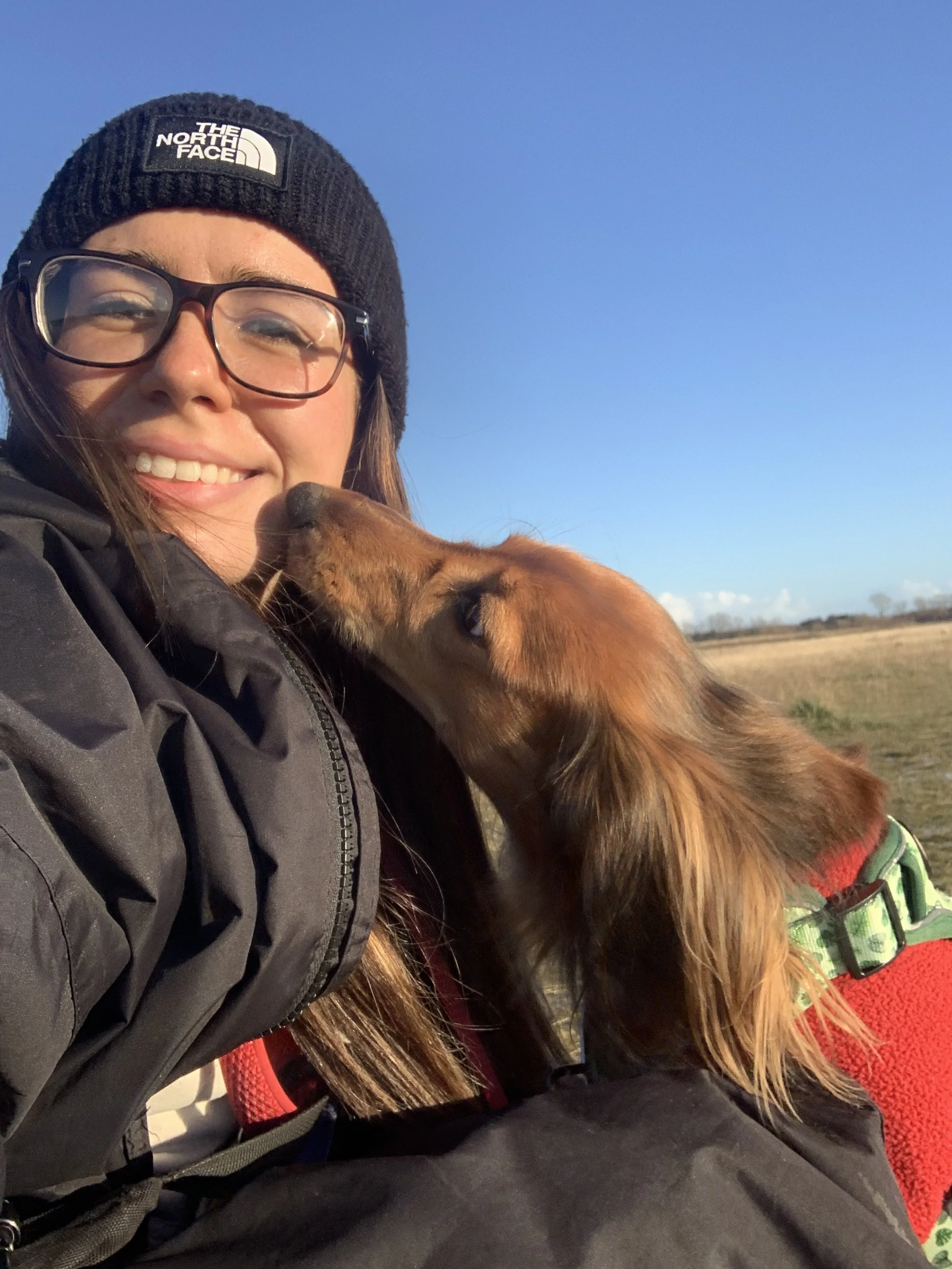 A woman with glasses smiling as a small brown dog with long ears kisses her face outdoors on a sunny day.   dog walker.