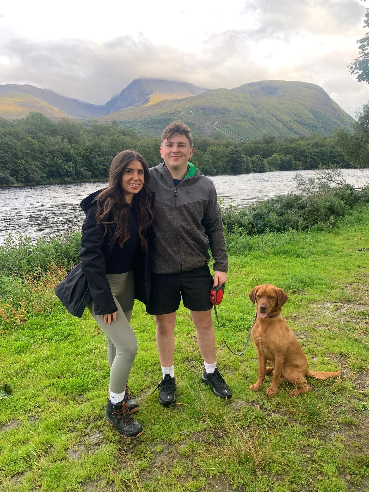 A smiling young woman and man standing side by side outdoors near a river, with a dog sitting on the grass next to them. The background features green hills and mountains under cloudy skies.