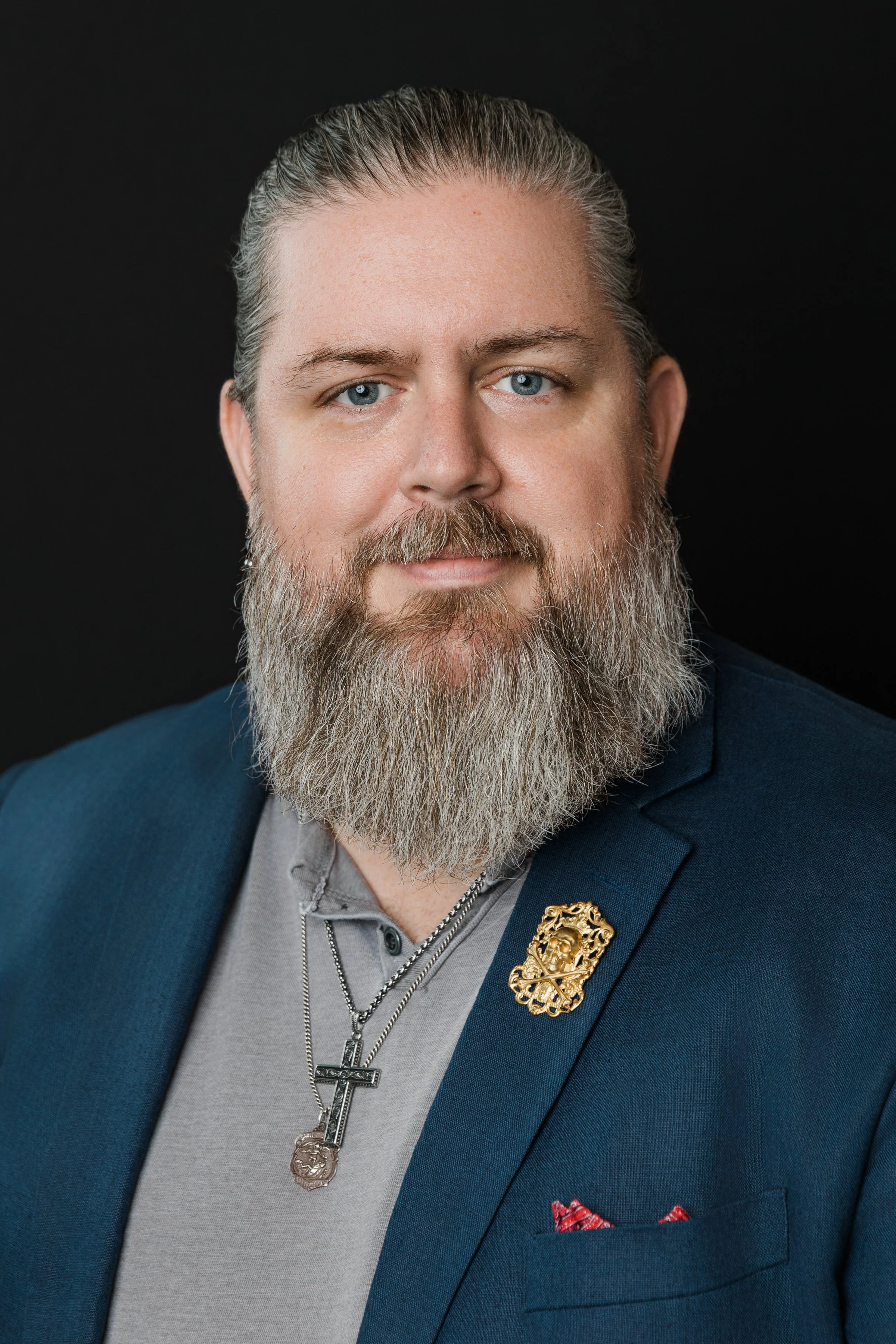 Sean McDaniel the Concierge of Strategic Relationships with a full gray beard and short gray hair, wearing a navy blazer, gray shirt, and layered necklaces with crosses, posing against a black background.