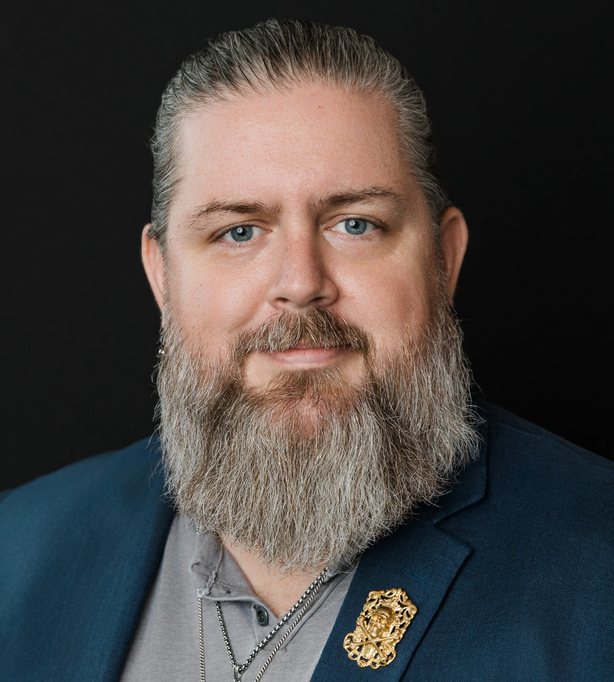 Close-up portrait of a man with a full beard, blue eyes, wearing a navy blazer, gray shirt, and a gold decorative pin, against a black background.