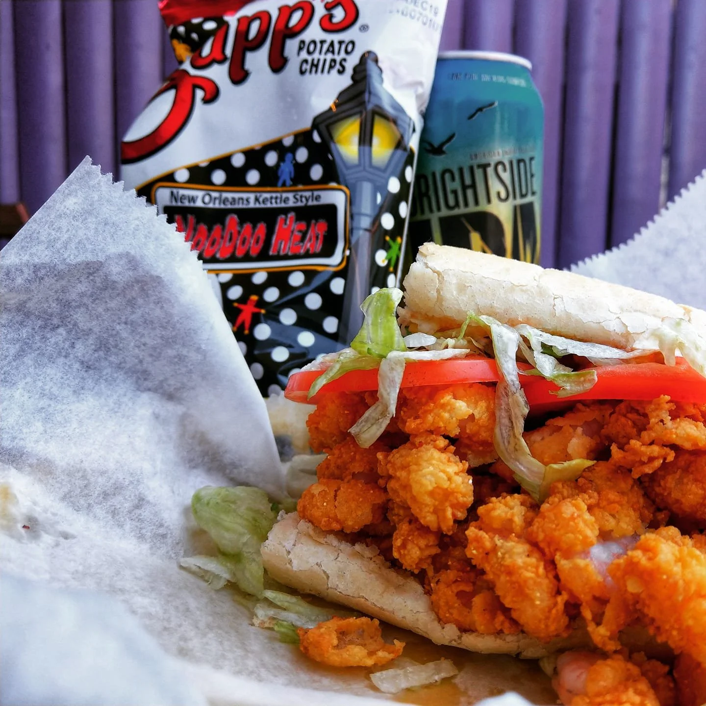 Fried chicken sandwich with lettuce, tomato, and mayonnaise in a bun, with a bag of potato chips and a can of Brightside beer in the background.