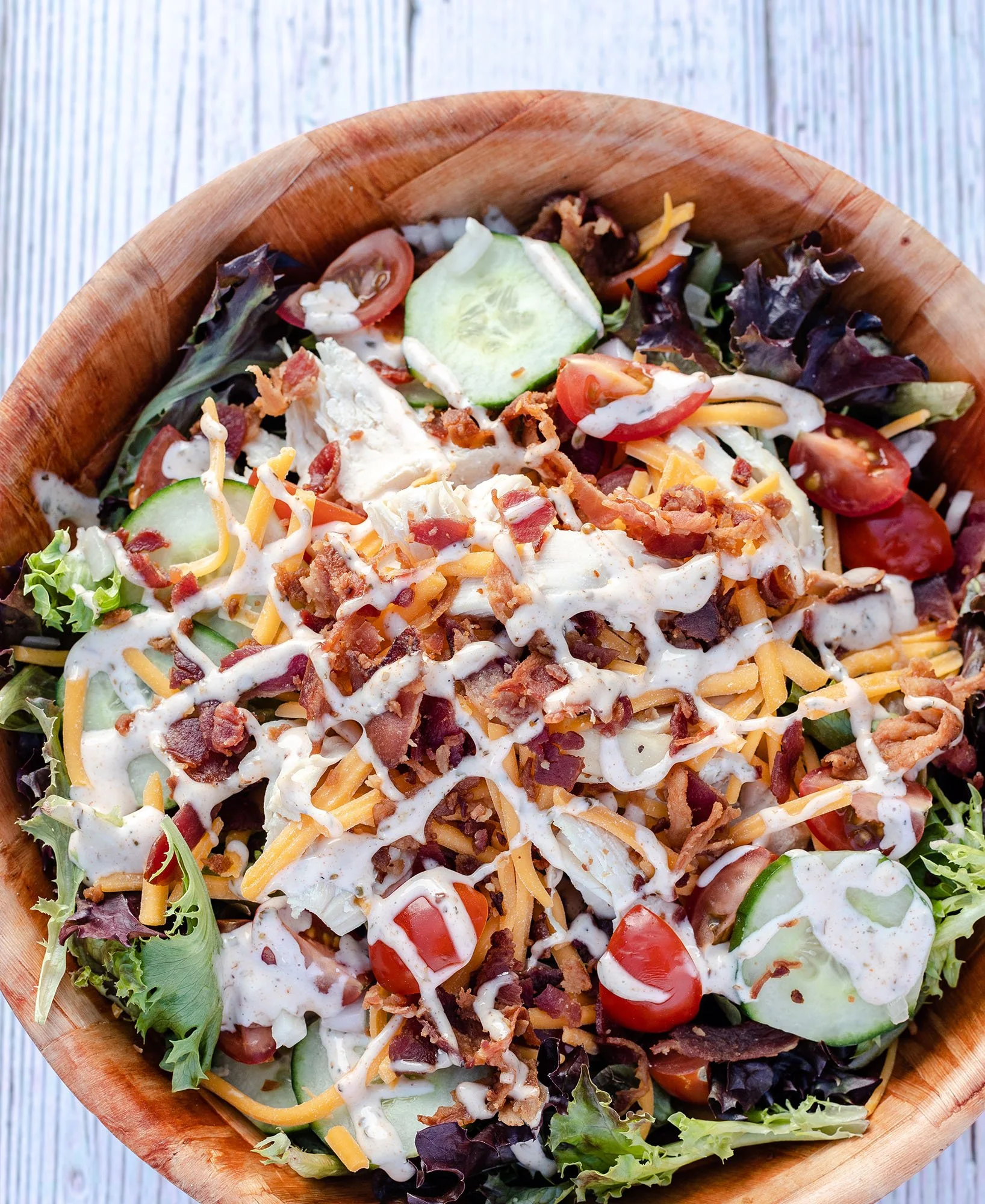 A bowl of mixed salad with cherry tomatoes, cucumber slices, shredded cheese, bacon bits, shredded chicken, shredded cheese, lettuce, and ranch dressing.