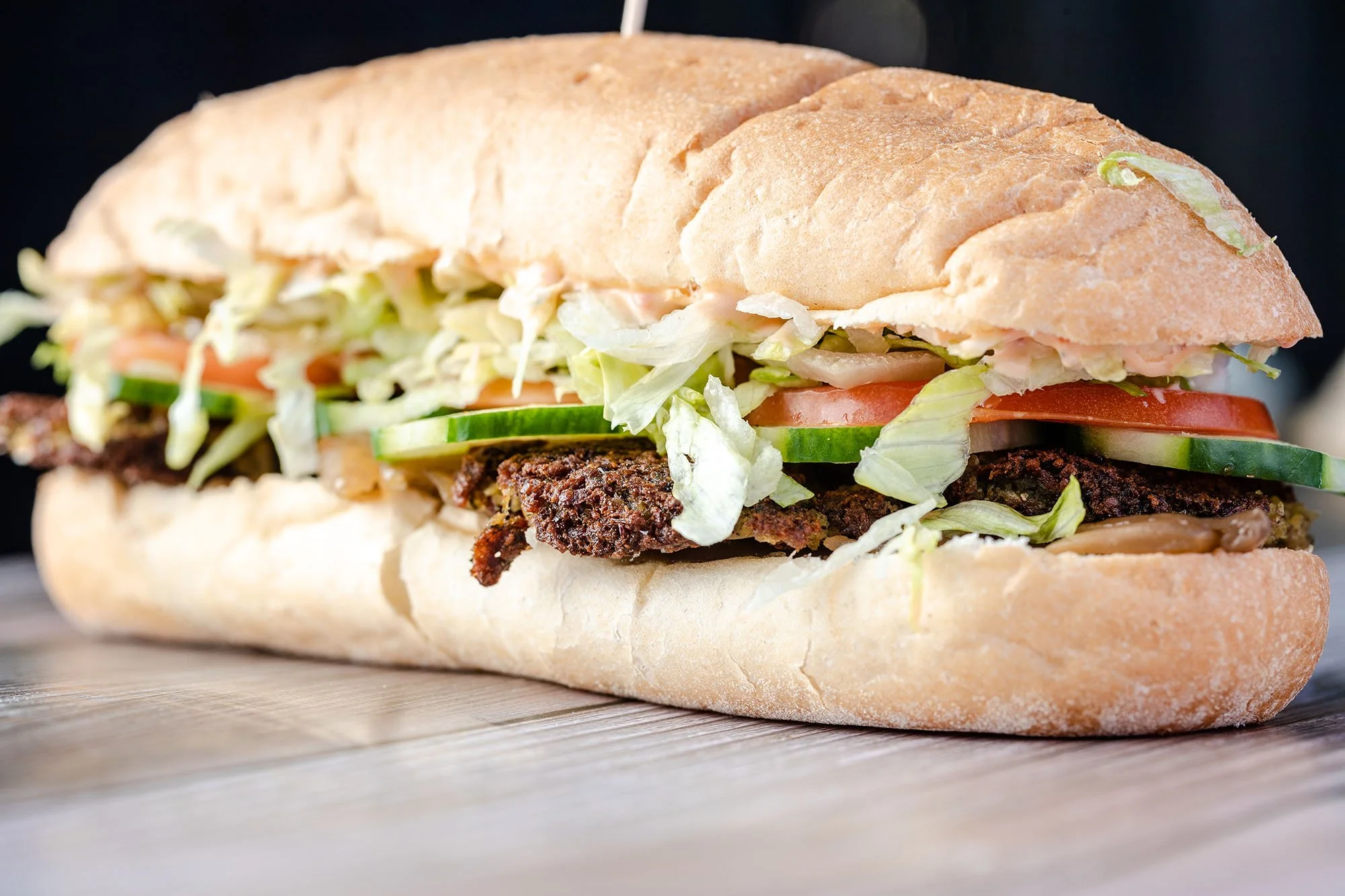A close-up of a po'boy sandwich with lettuce, tomato, cucumber, grilled meat, on a French bread roll, on a wooden surface.
