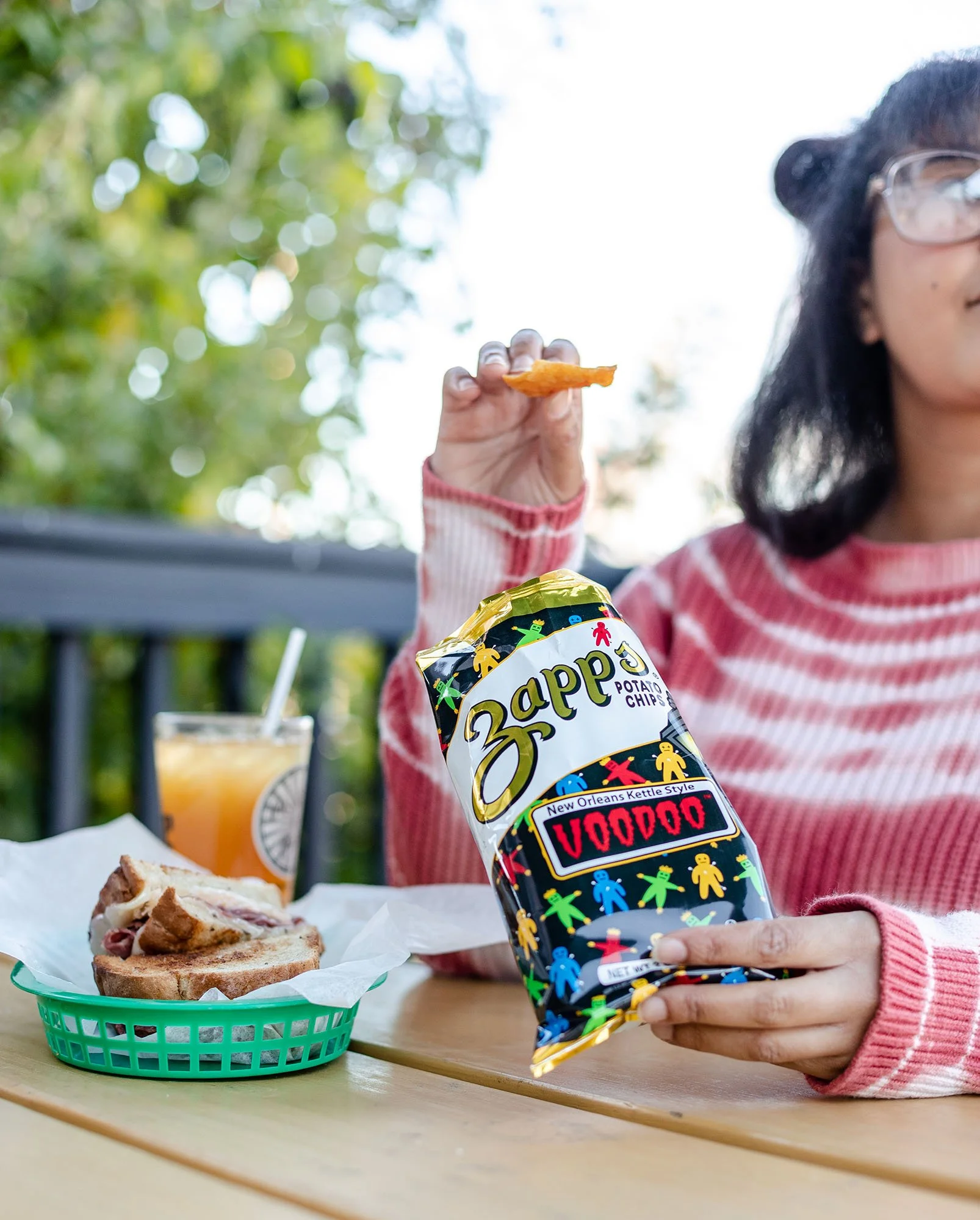 A person holding a bag of Zapp's potato chips, with a sandwich and a drink on the table in front of them, sitting outside with trees in the background.