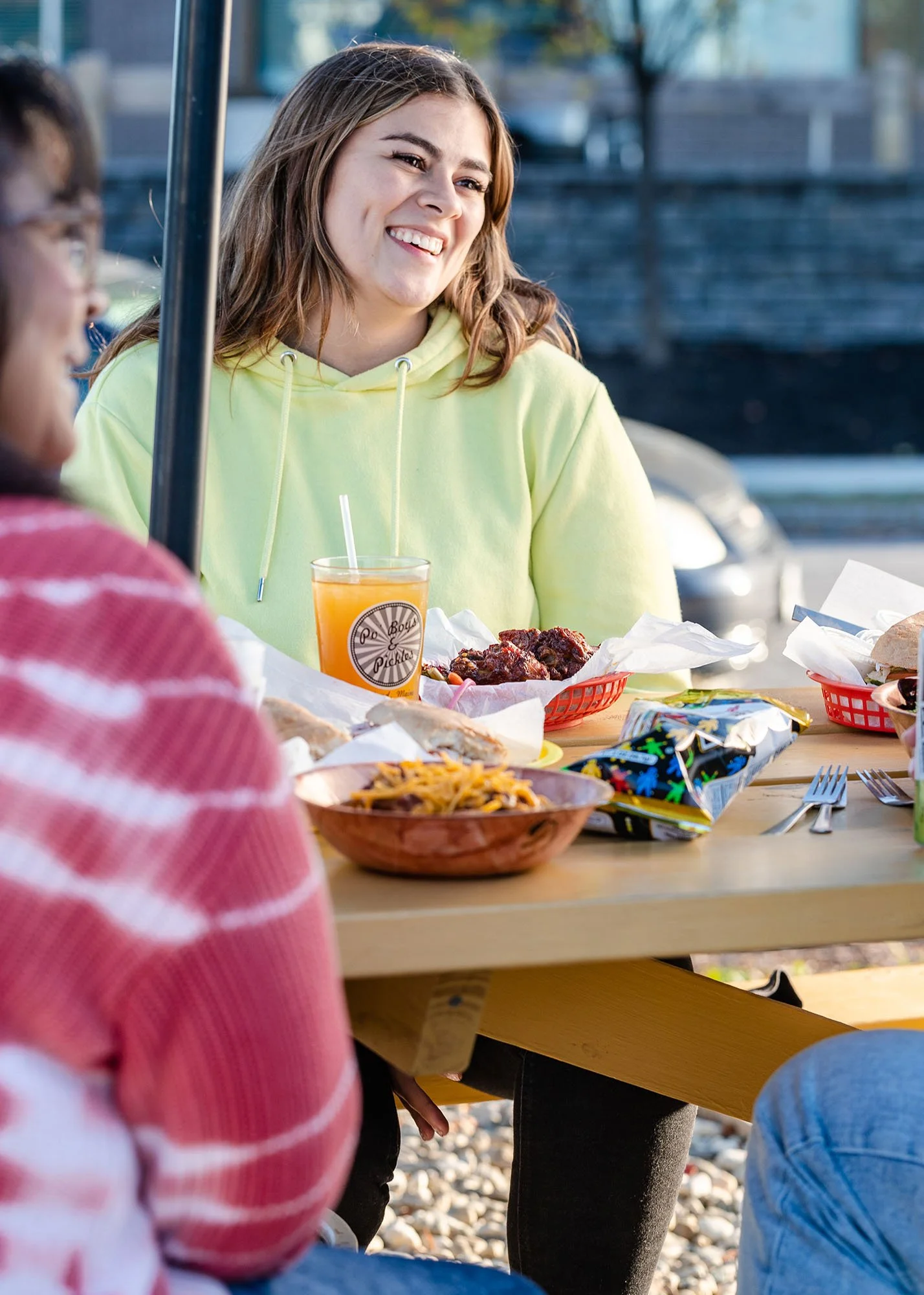 A group of people enjoying a meal outdoors at a picnic table, with food and drinks, amid a casual social gathering.