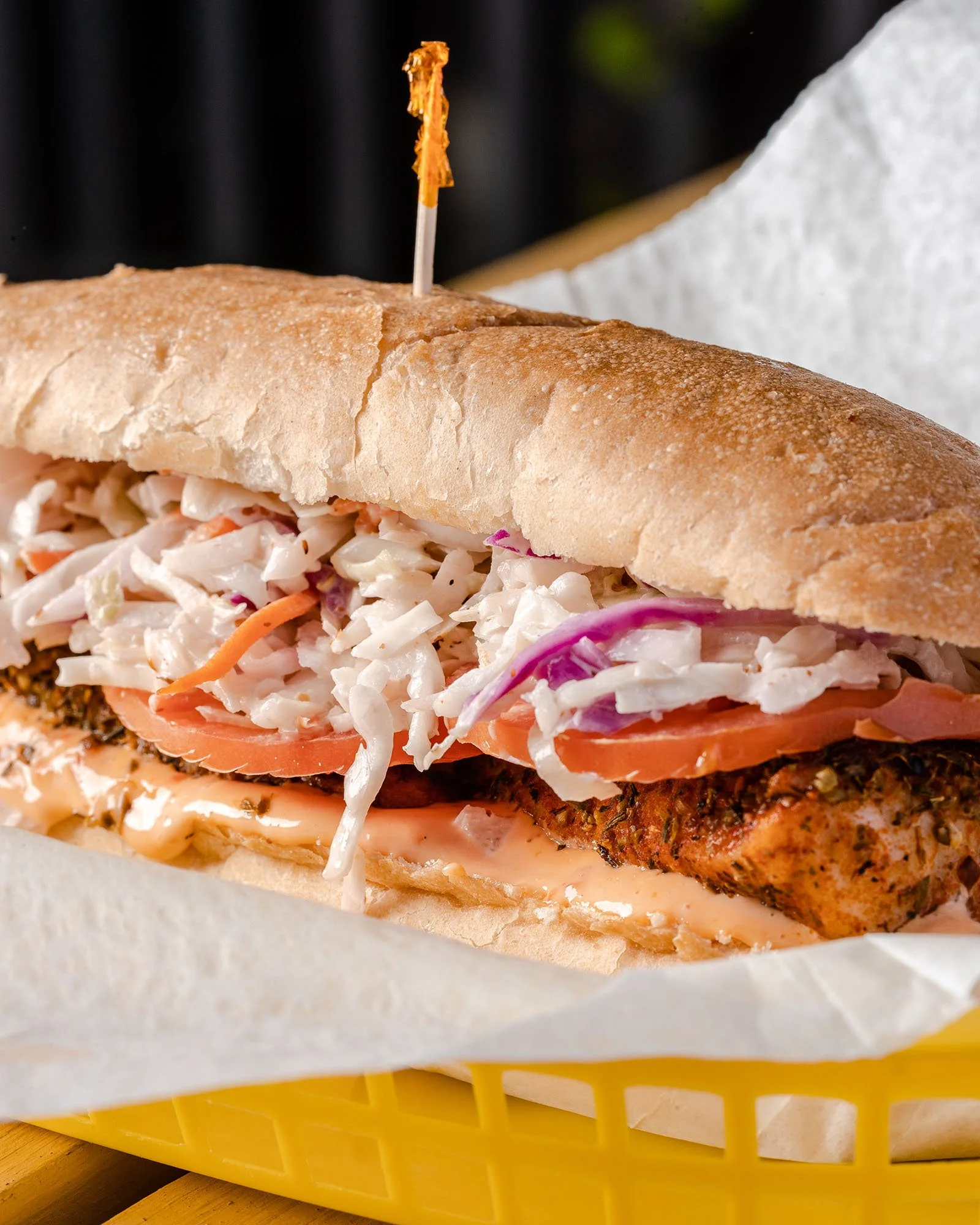 Close-up of a po'boy sandwich with coleslaw, tomato slices, grilled meat, and sauce, topped with a toothpick