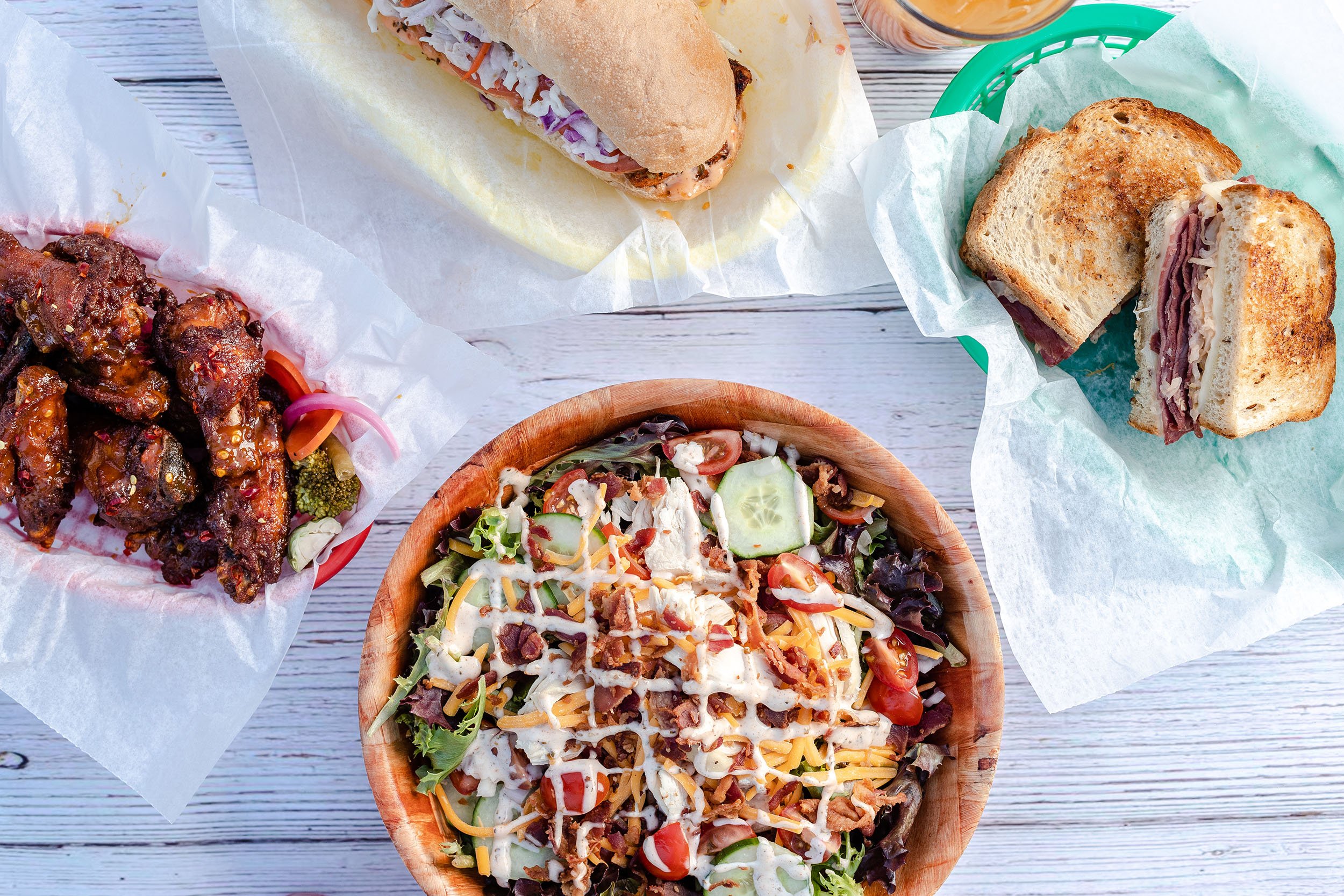 A top-down view of a meal including a bowl of salad, two sandwiches with bread and various fillings, a serving of buffalo chicken wings with sauce and pickled vegetables, all arranged on a white wooden surface.