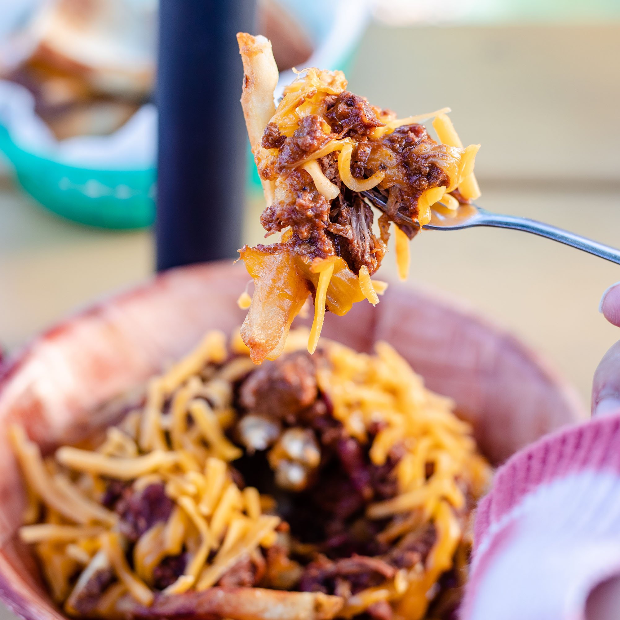 A fork with a bite of chili, cheese, and sour cream above a bowl of chili with shredded cheese and sour cream.