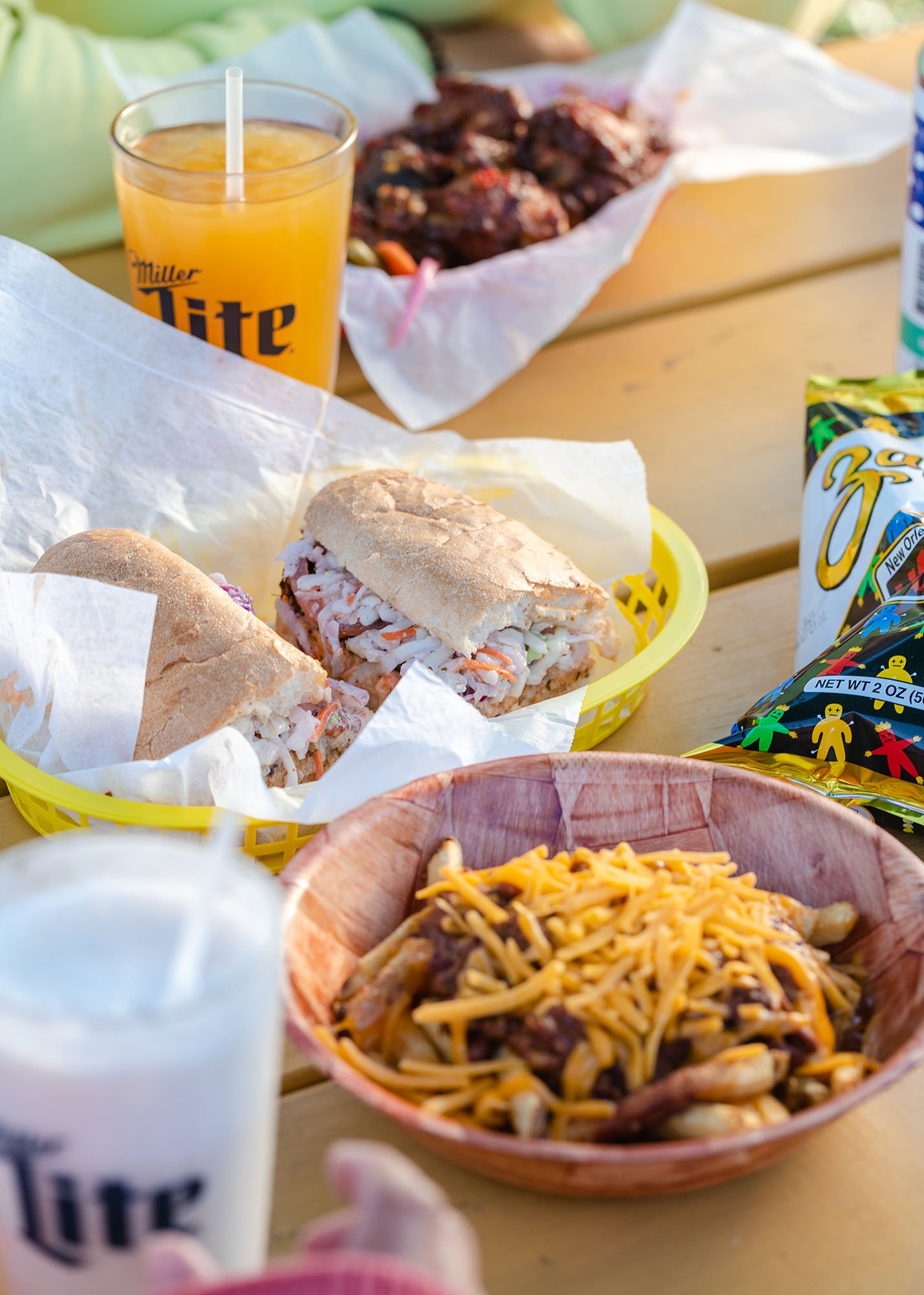 A picnic table with a basket of pulled pork sandwiches, a bowl of chili with shredded cheese, a glass of orange juice, a tray of cooked chicken wings, and snack bags of chips.